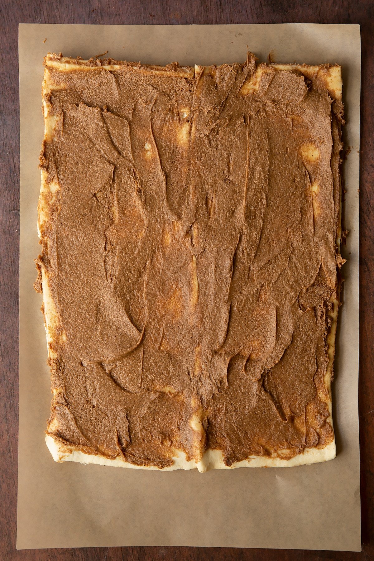 Two strips of unrolled cinnamon swirl dough on a piece of brown baking paper. The cinnamon butter filling is spread evenly.