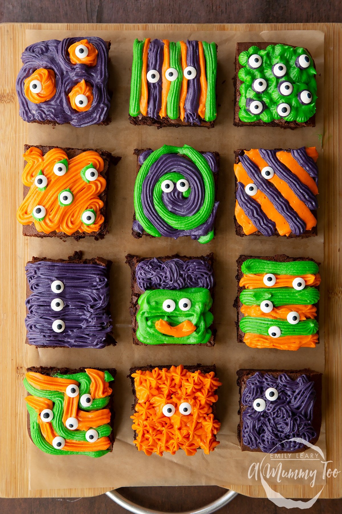 12 Halloween brownies arranged on a board lined with baking paper. The brownies are decorated with purple, green and orange frosting and candy eyes.