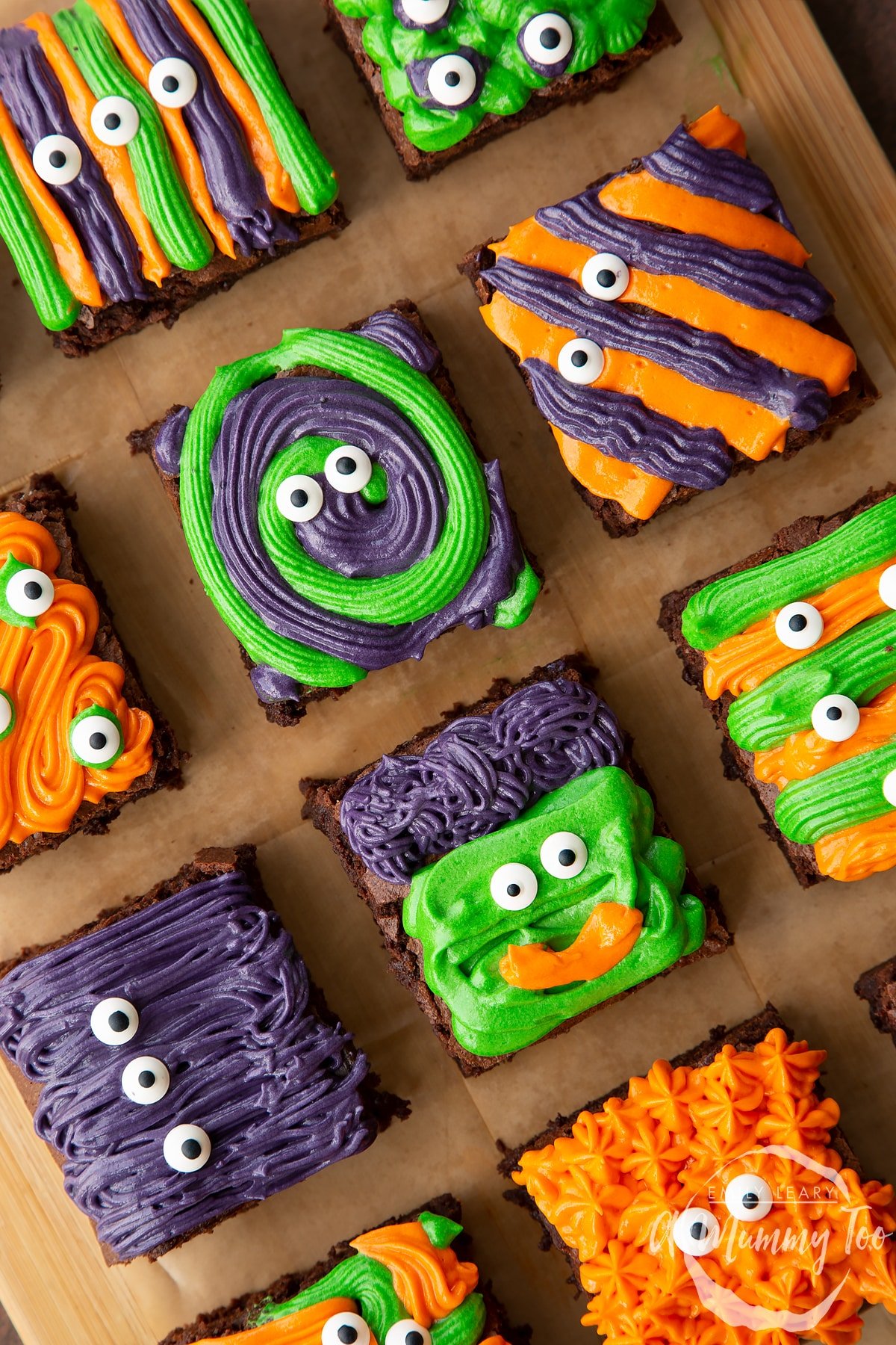 Halloween brownies arranged on a board lined with baking paper. The brownies are decorated with purple, green and orange frosting and candy eyes.