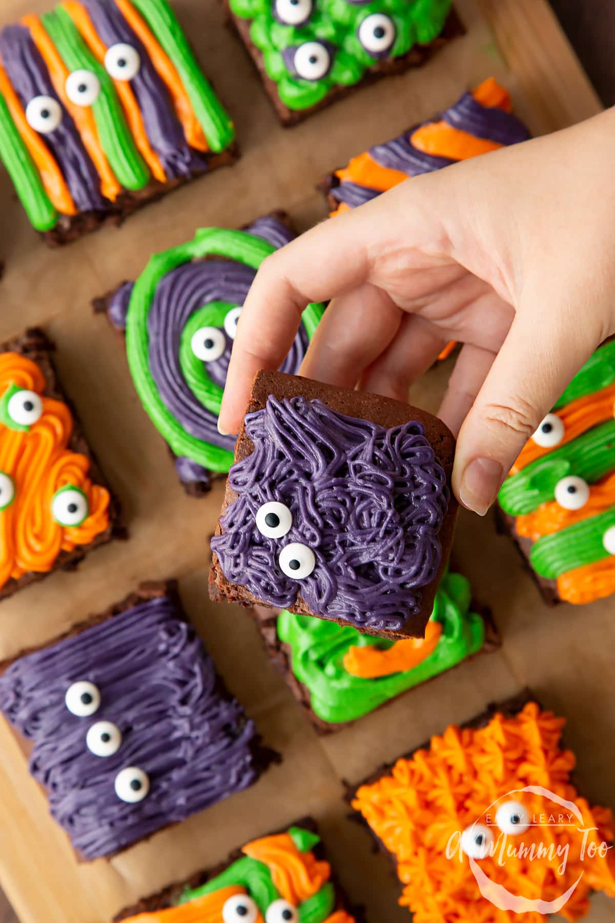 Halloween brownies arranged on a board lined with baking paper. A hand holds a purple frosted brownie with candy eyes.