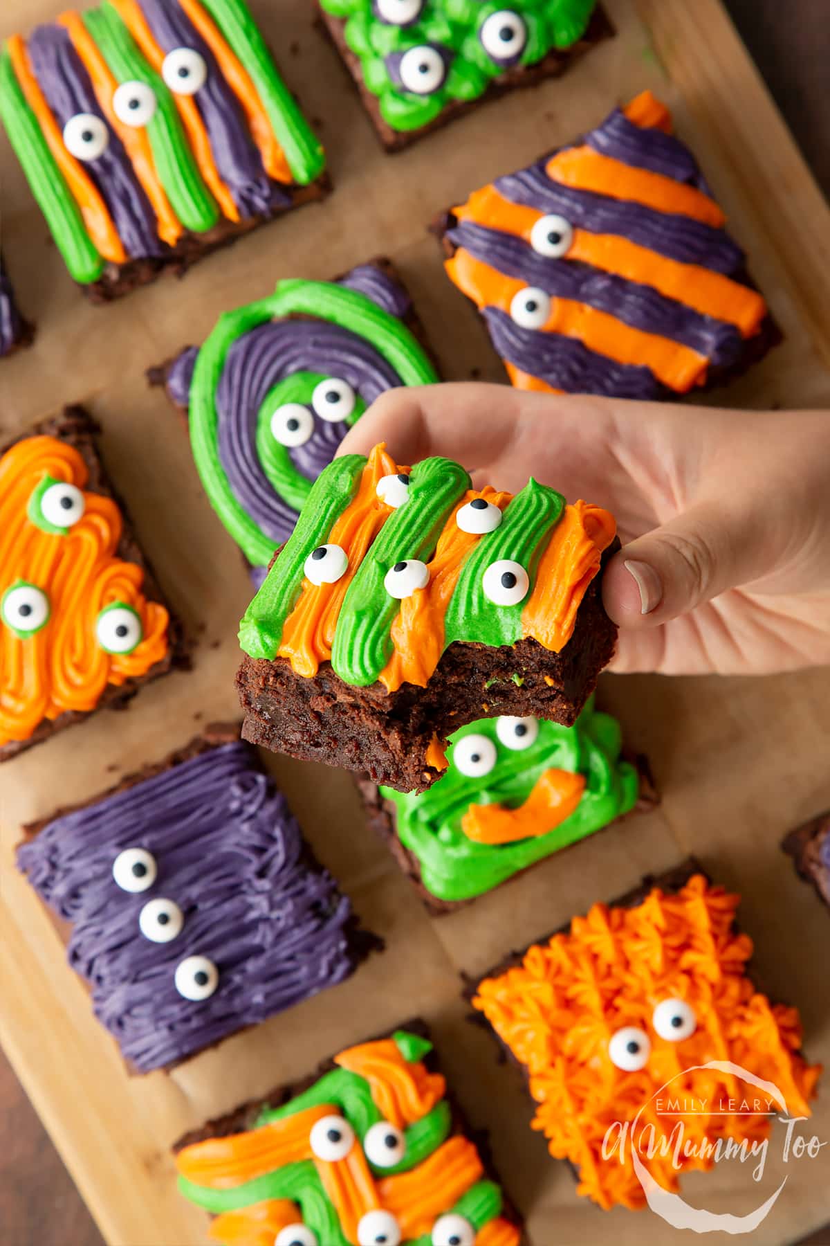 Halloween brownies arranged on a board lined with baking paper. A hand holds a purple and green frosted brownie with candy eyes.