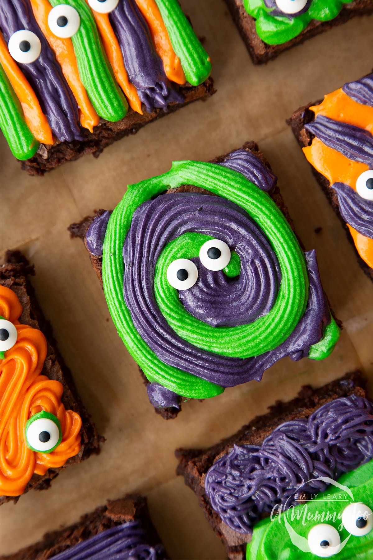 Close up of a purple and green frosted Halloween brownie with candy eyes, resting on a board lined with baking paper.
