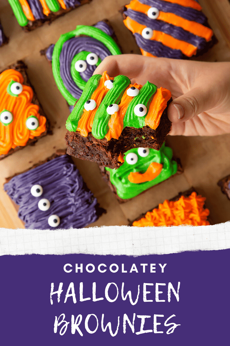 Halloween brownies arranged on a board lined with baking paper. A hand holds a orange and green frosted one. Caption reads: chocolatey Halloween brownies