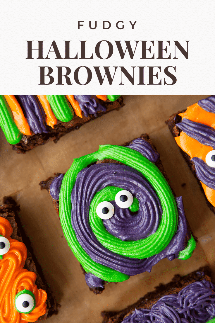 Close up of a purple and green frosted Halloween brownie with candy eyes, resting on a board lined with baking paper. Caption reads: fudgy Halloween brownies