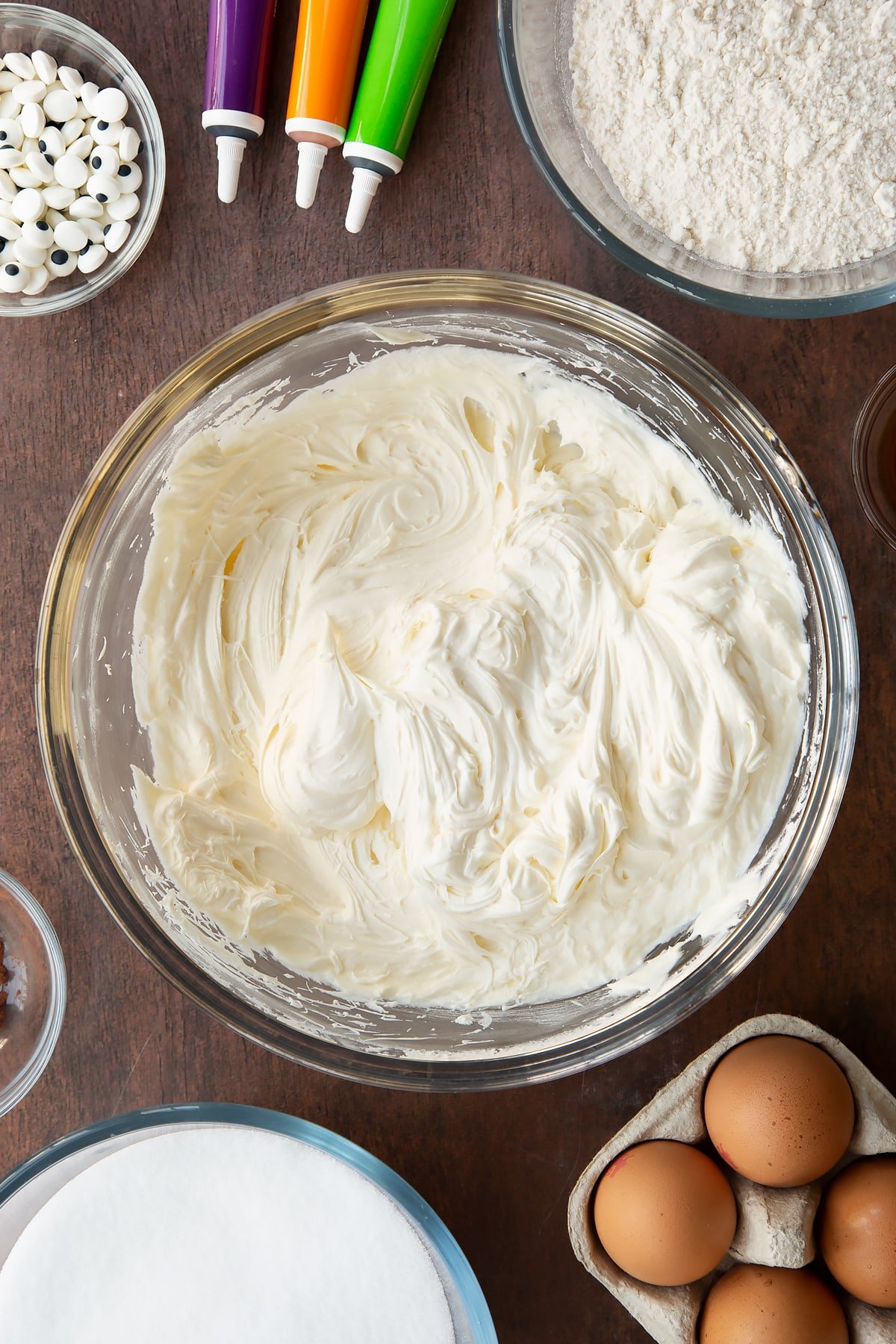 Cream cheese frosting in a glass bowl. Ingredients to make Halloween brownies surround the bowl.