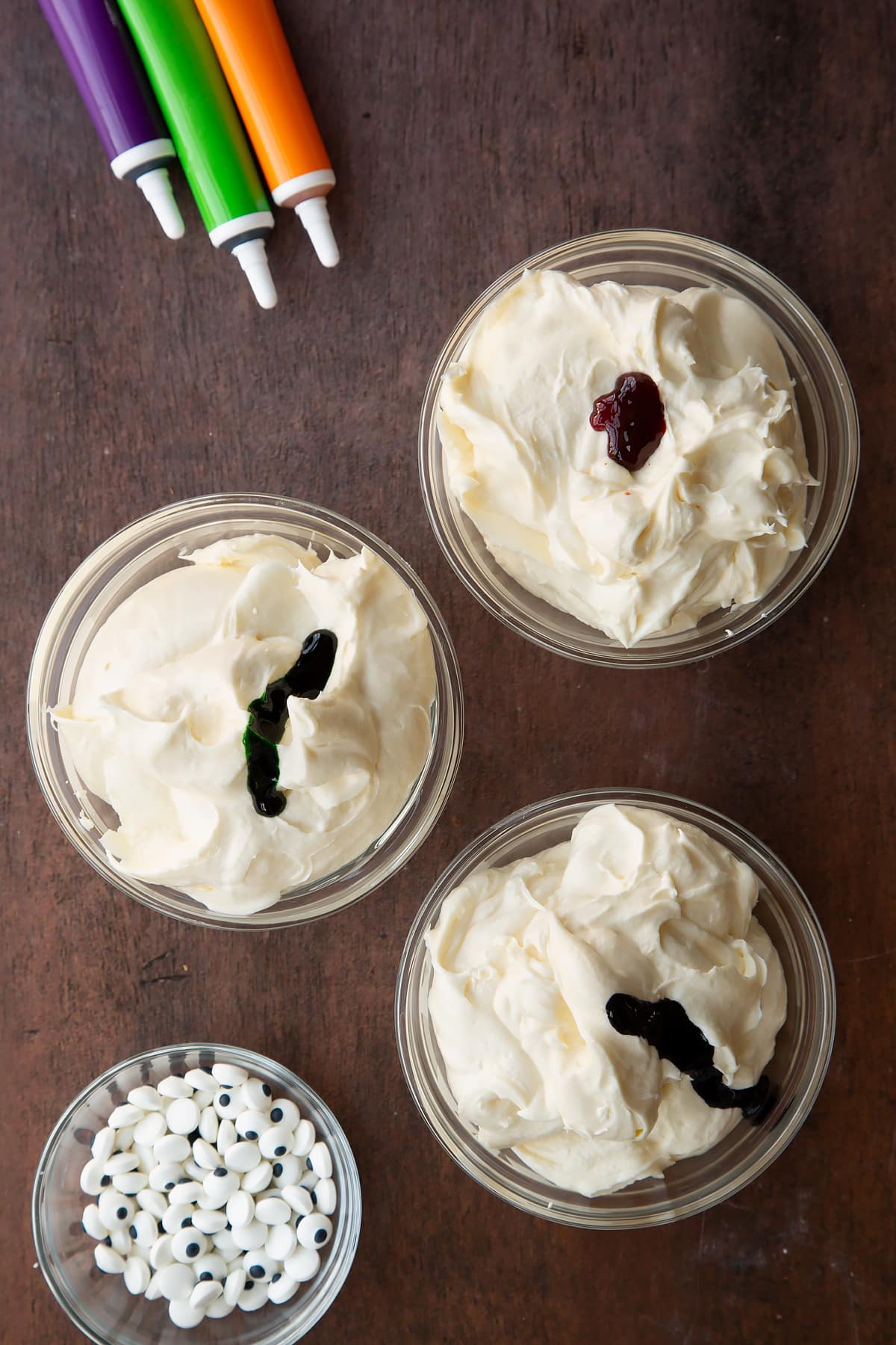 Cream cheese frosting in three glass bowls. Each has food colouring on top - orange, green or purple.