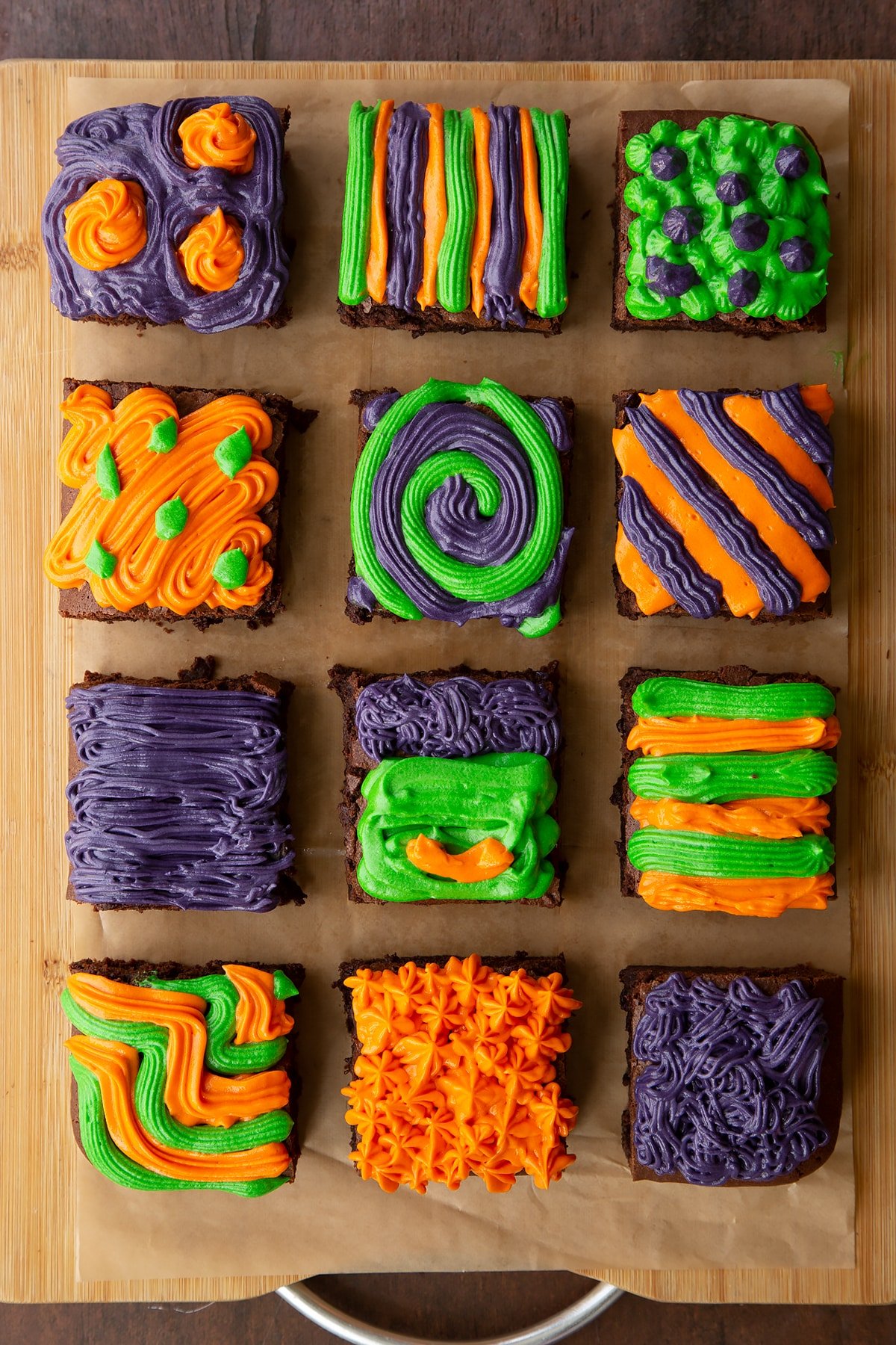 Halloween brownie on baking paper, cut into 12 pieces. Orange, green and purple frosting has been piped on top of the brownies in various patterns.