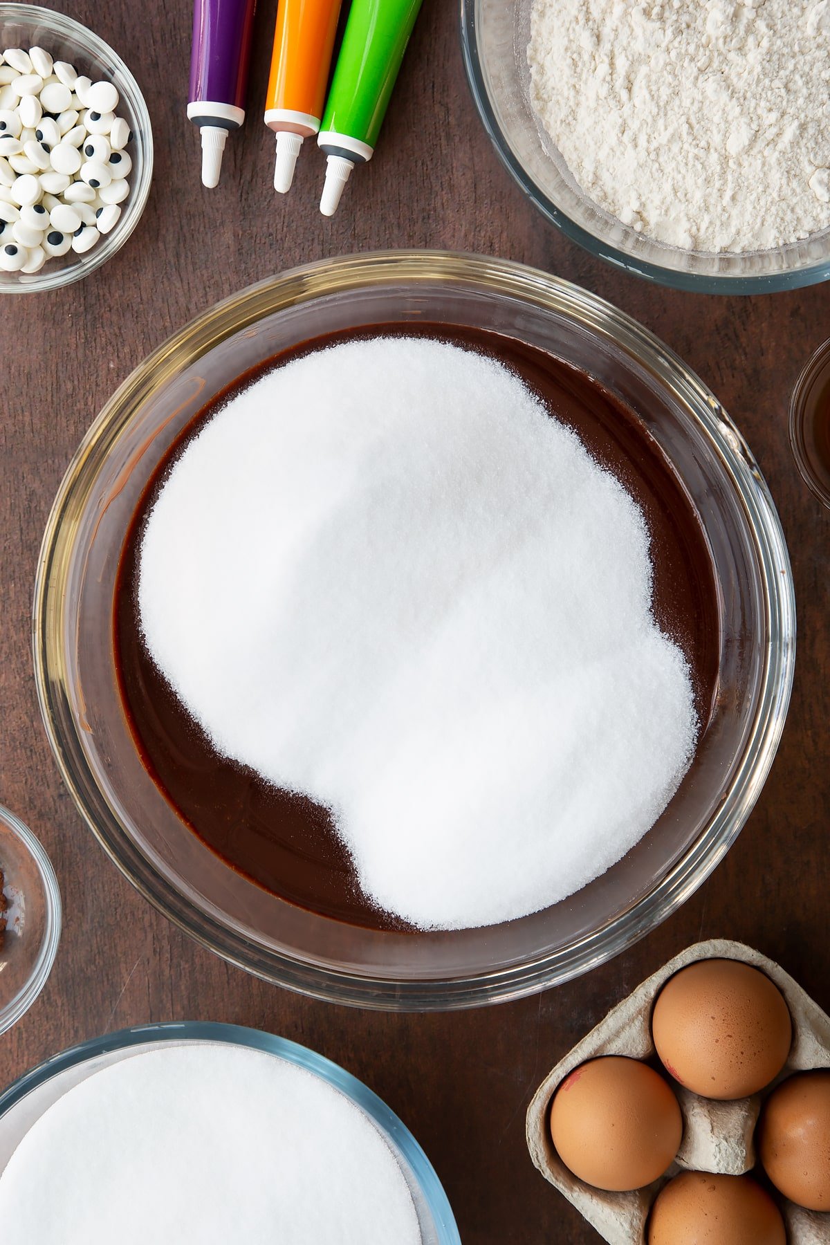 Melted butter and dark chocolate in a glass bowl with sugar on top. Ingredients to make Halloween brownies surround the bowl.