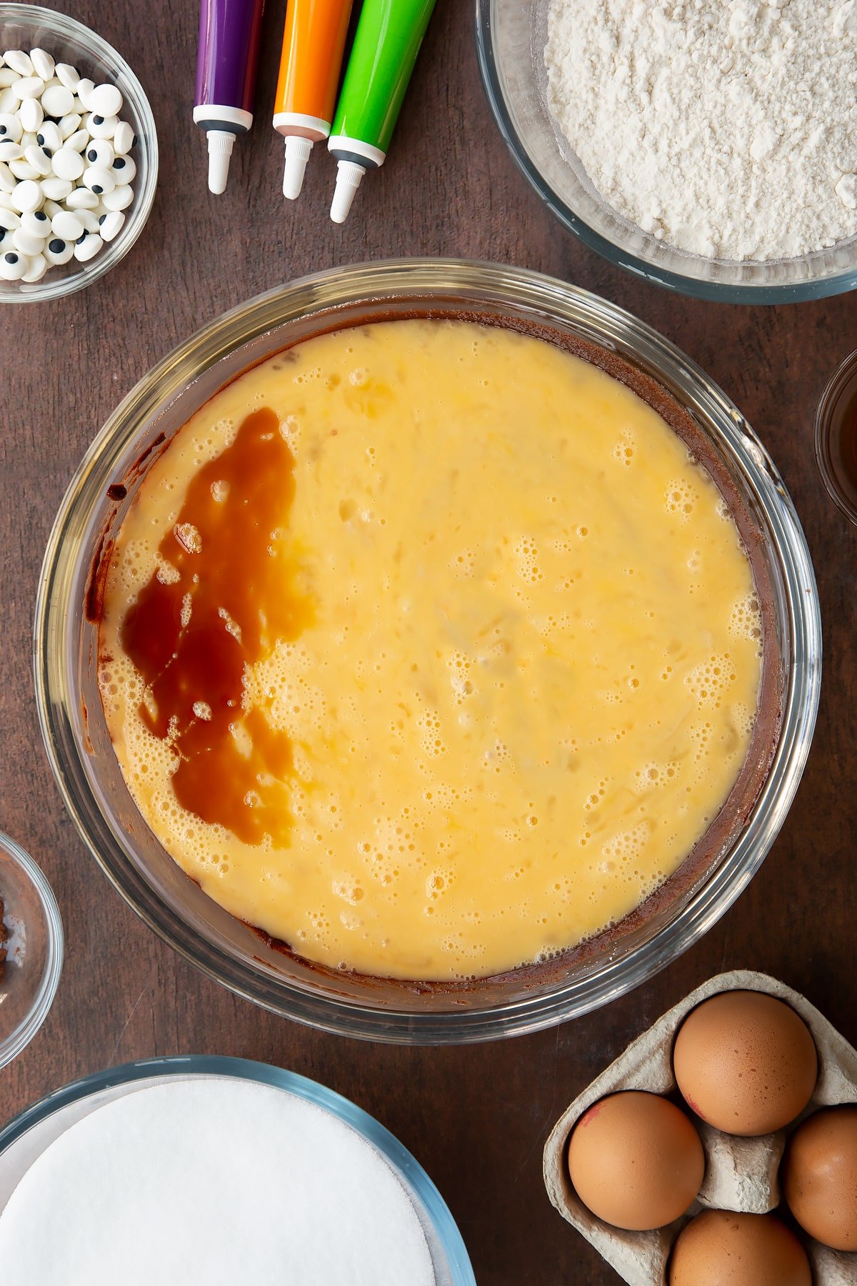 Melted butter, dark chocolate and sugar in a glass bowl with beaten eggs and vanilla on top. Ingredients to make Halloween brownies surround the bowl.