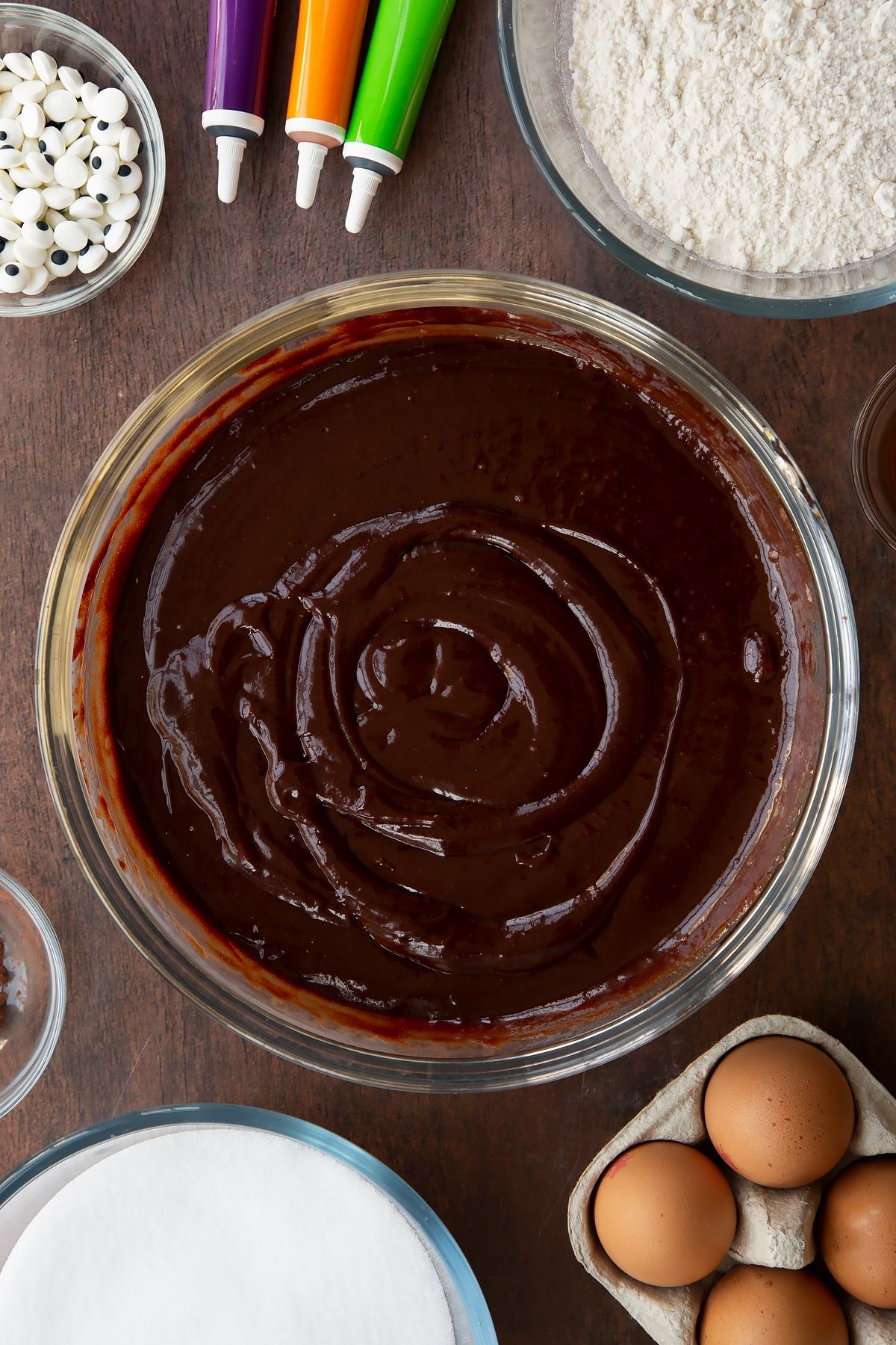 Melted butter, dark chocolate, eggs, vanilla and sugar in a glass bowl. Ingredients to make Halloween brownies surround the bowl.