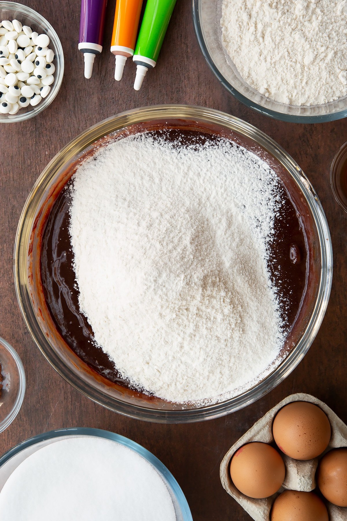 Melted butter, dark chocolate, eggs, vanilla and sugar in a glass bowl with flour on top. Ingredients to make Halloween brownies surround the bowl.