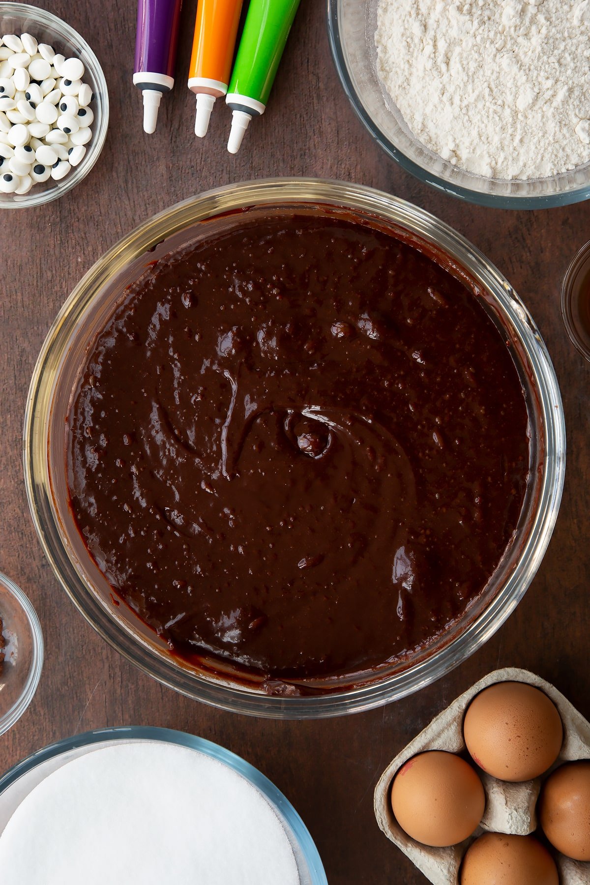 Brownie batter in a glass bowl. Ingredients to make Halloween brownies surround the bowl.