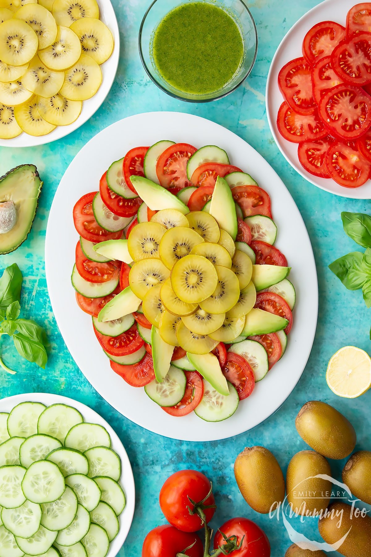 Sliced tomatoes, cucumber, avocado and golden kiwi on a white oval plate. Ingredients to make a kiwi feta salad surround the platter.