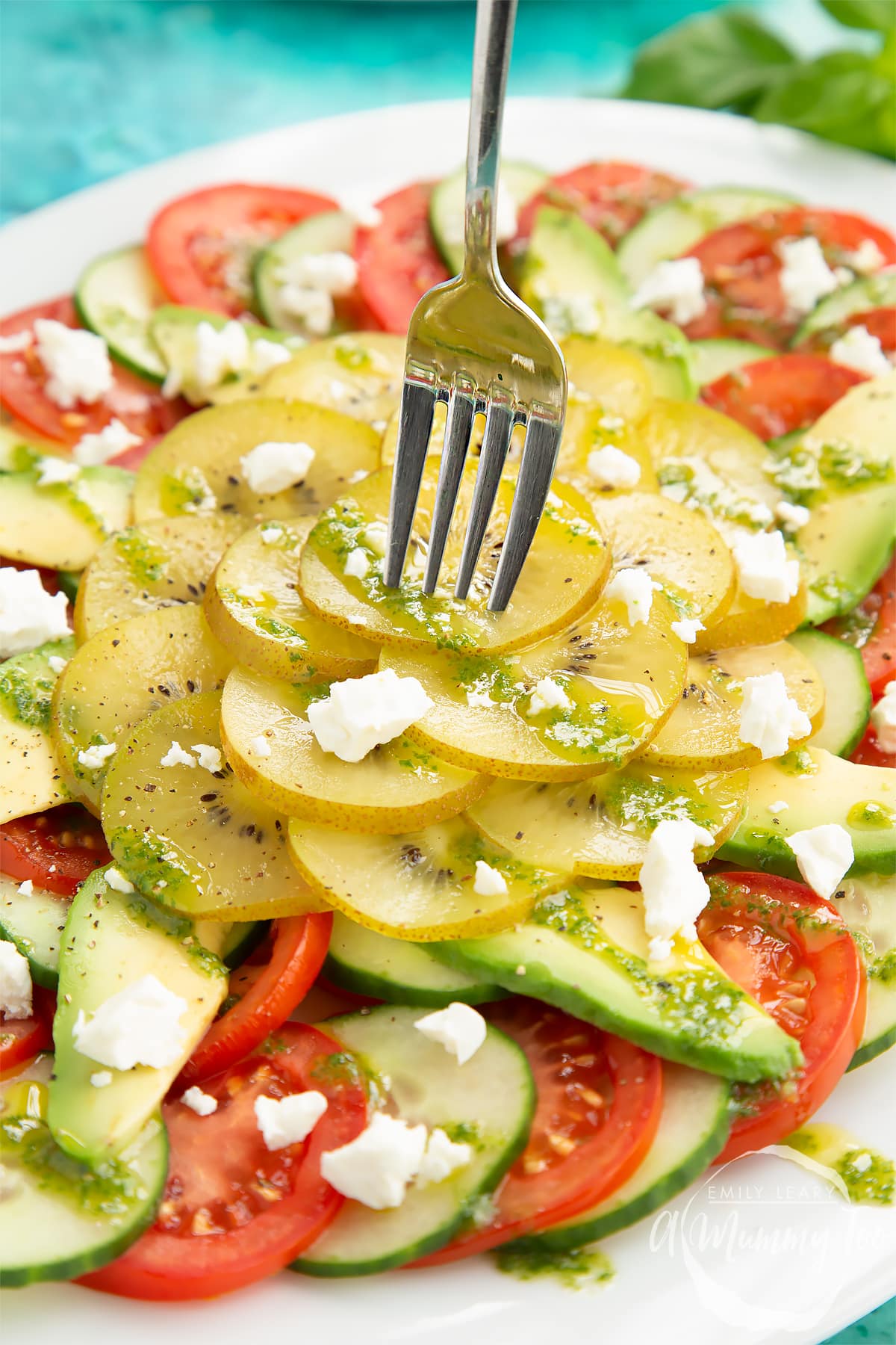 Close up of kiwi feta salad with basil oil on a white platter. A fork digs into the salad.