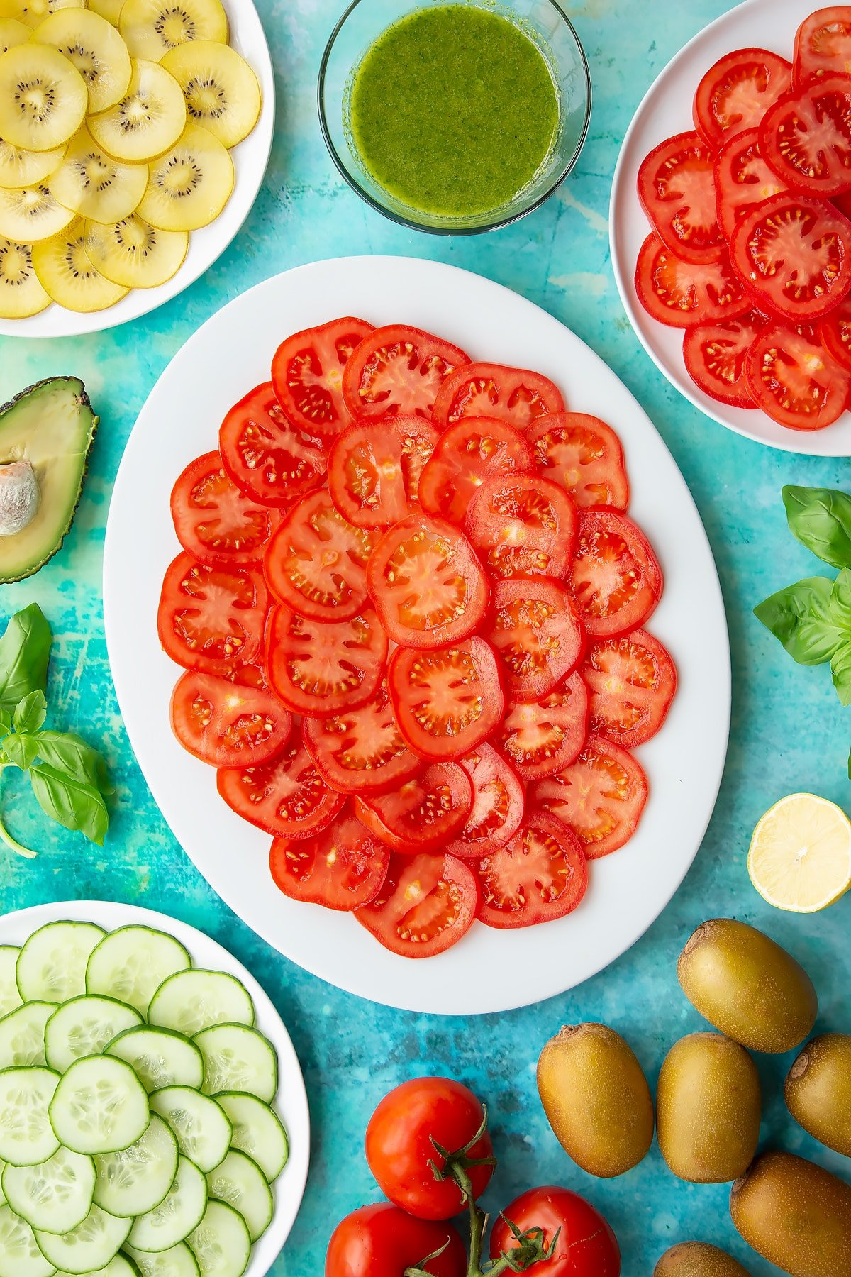 Sliced tomatoes on a white oval plate. Ingredients to make a kiwi feta salad surround the platter.