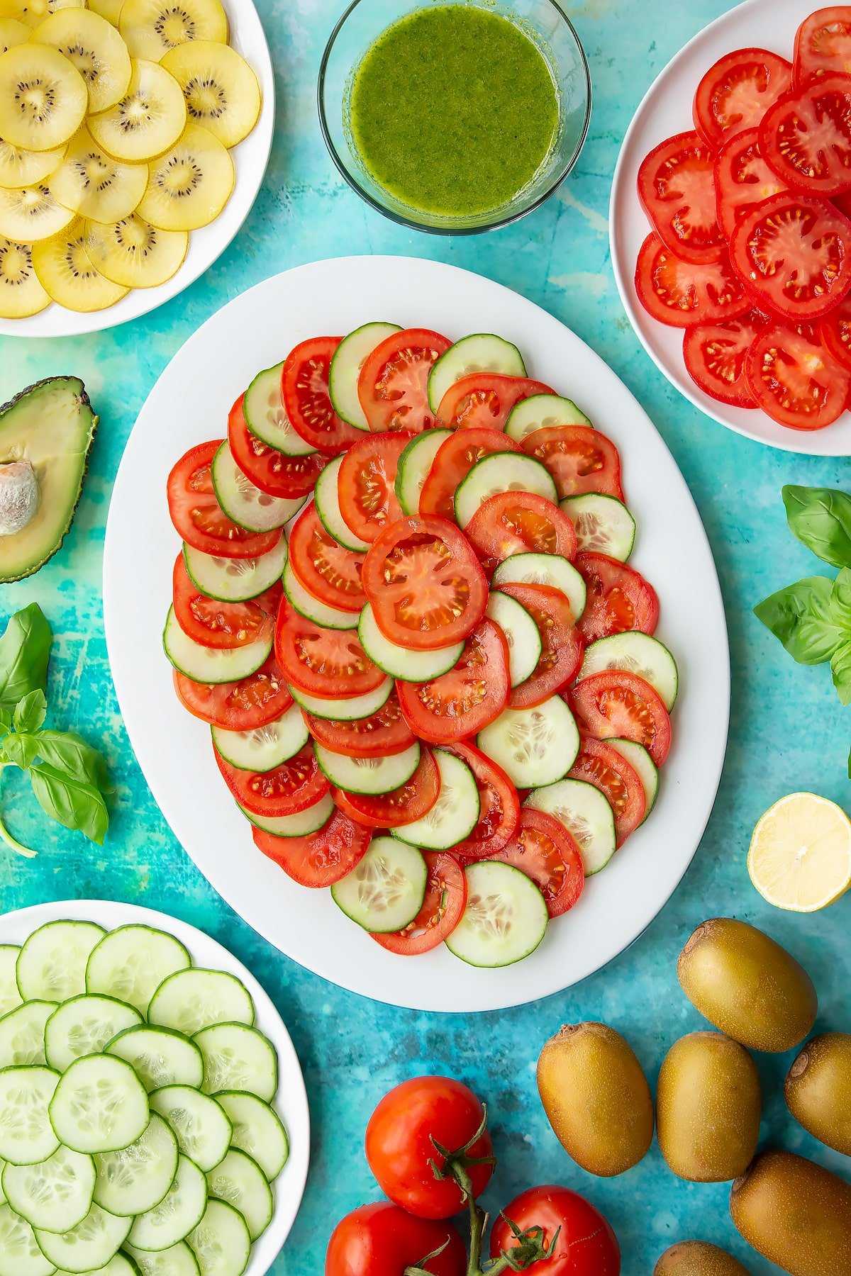 Sliced tomatoes and cucumber on a white oval plate. Ingredients to make a kiwi feta salad surround the platter.