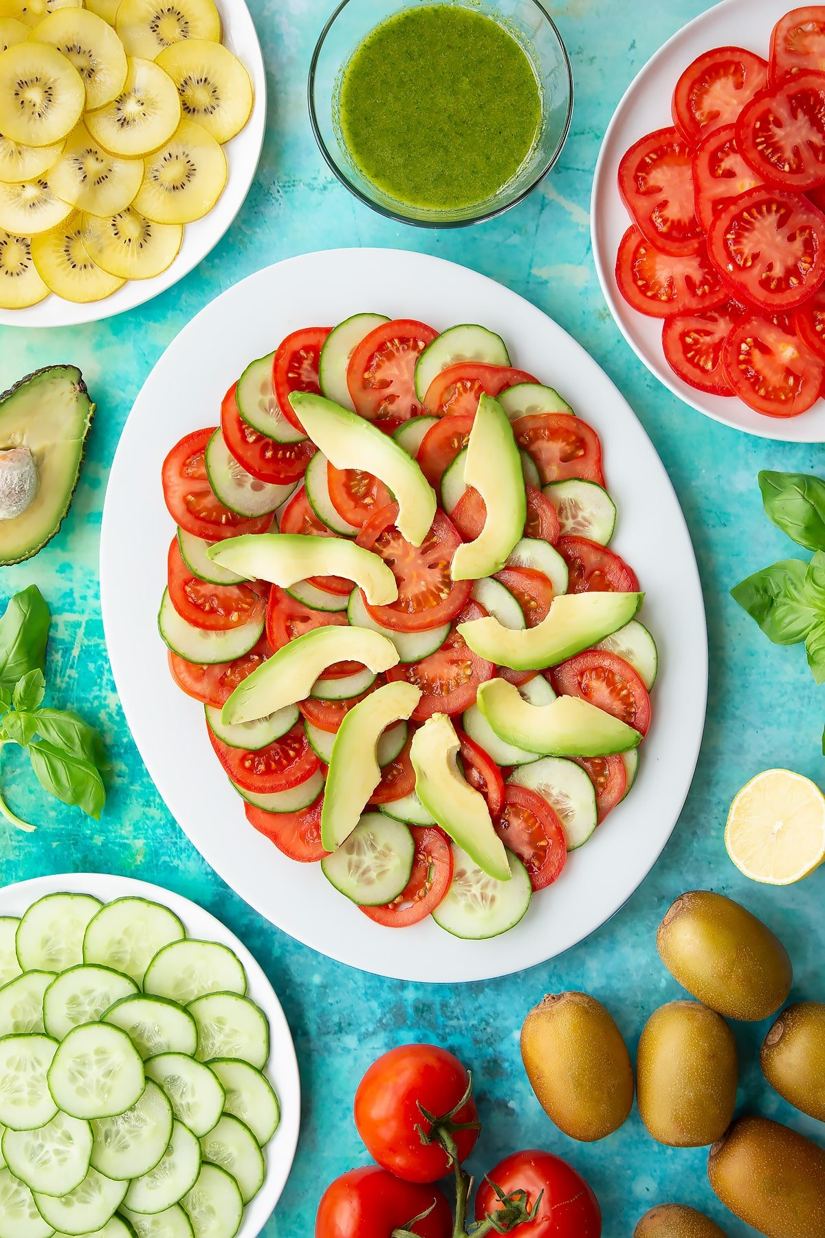 Sliced tomatoes, cucumber and avocado on a white oval plate. Ingredients to make a kiwi feta salad surround the platter.