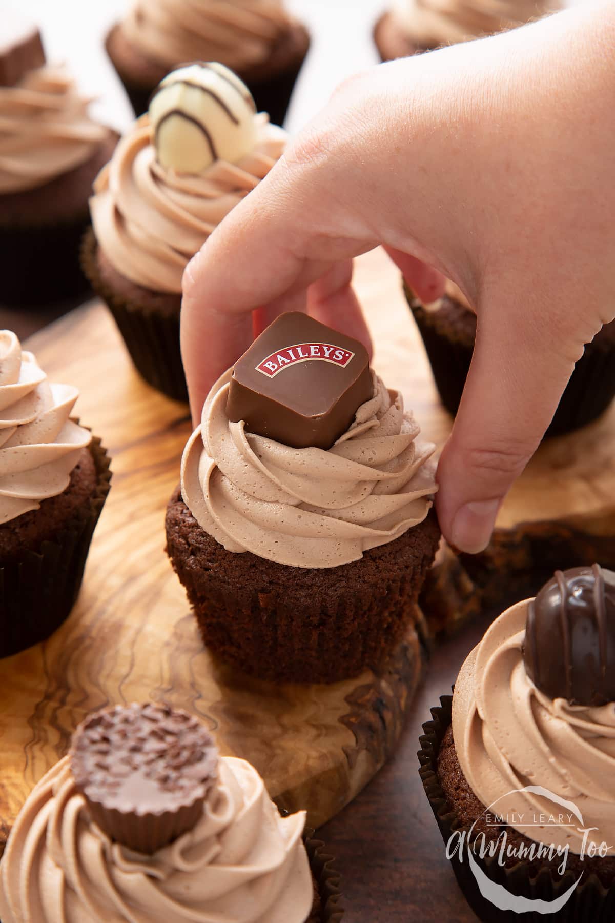 Baileys chocolate cupcakes topped with Baileys icing and a Baileys chocolate. A hand reaches for one.