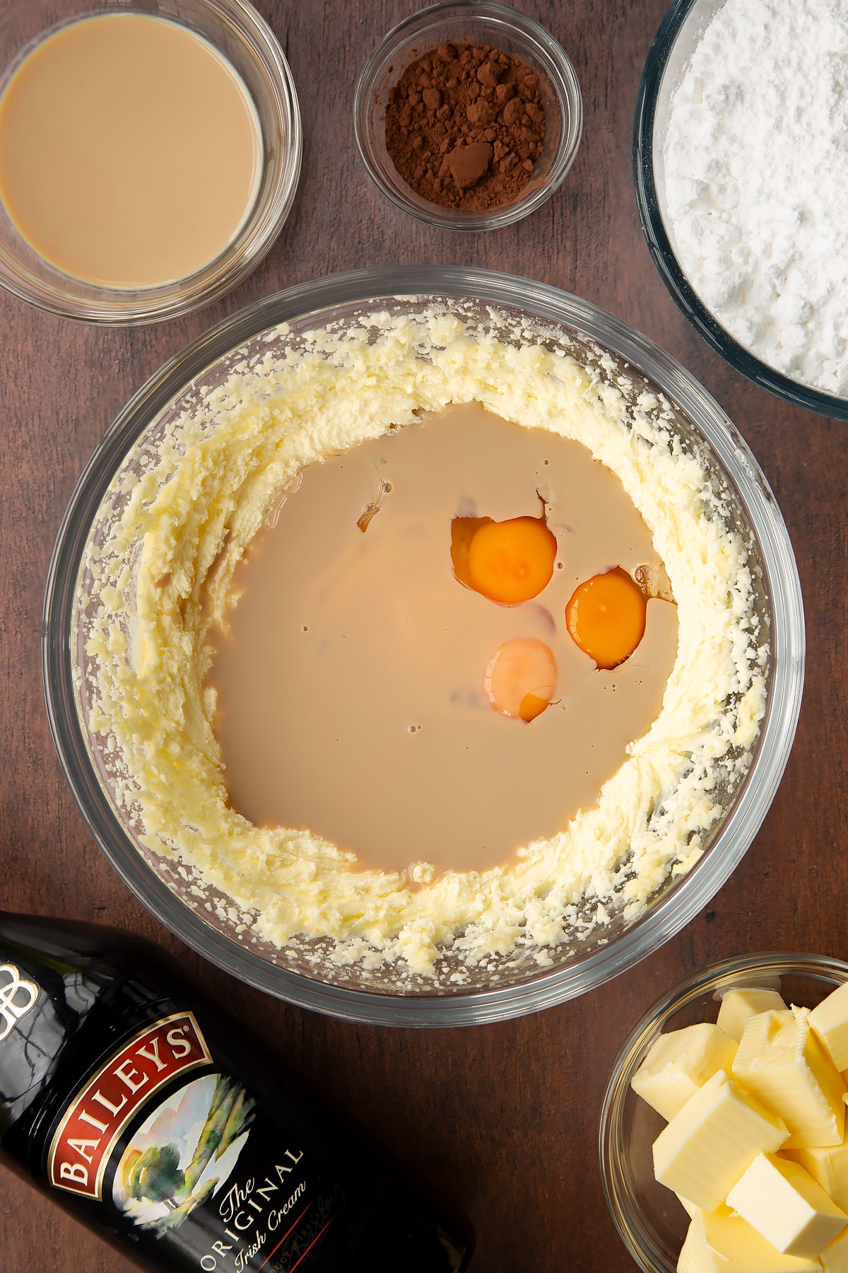 Butter, oil and sugar, whisked together in a glass mixing bowl with Baileys and eggs on top. Ingredients to make Baileys chocolate cupcakes surround the bowl.