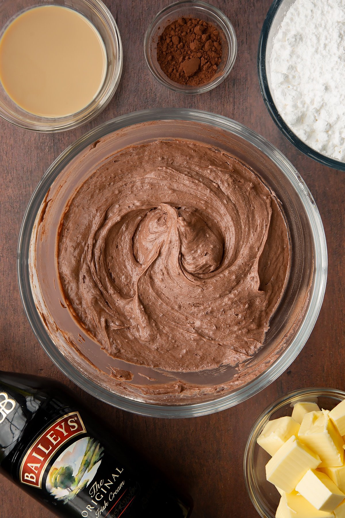 Baileys chocolate cupcake batter in a glass mixing bowl. Ingredients to make Baileys chocolate cupcakes surround the bowl.