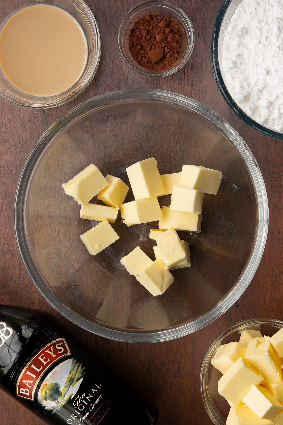 Cubed butter in a glass mixing bowl. Ingredients to make Baileys chocolate cupcakes surround the bowl.