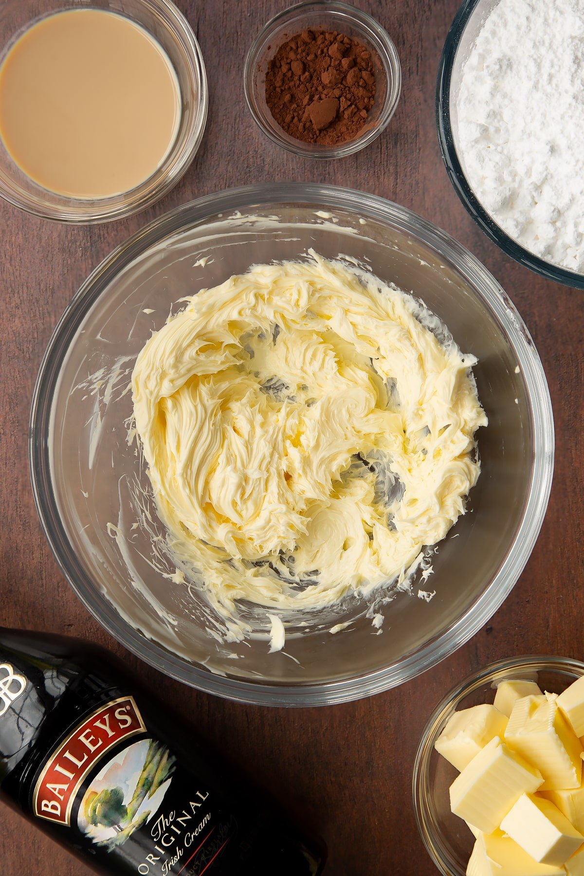 Whipped butter in a glass mixing bowl. Ingredients to make Baileys chocolate cupcakes surround the bowl.