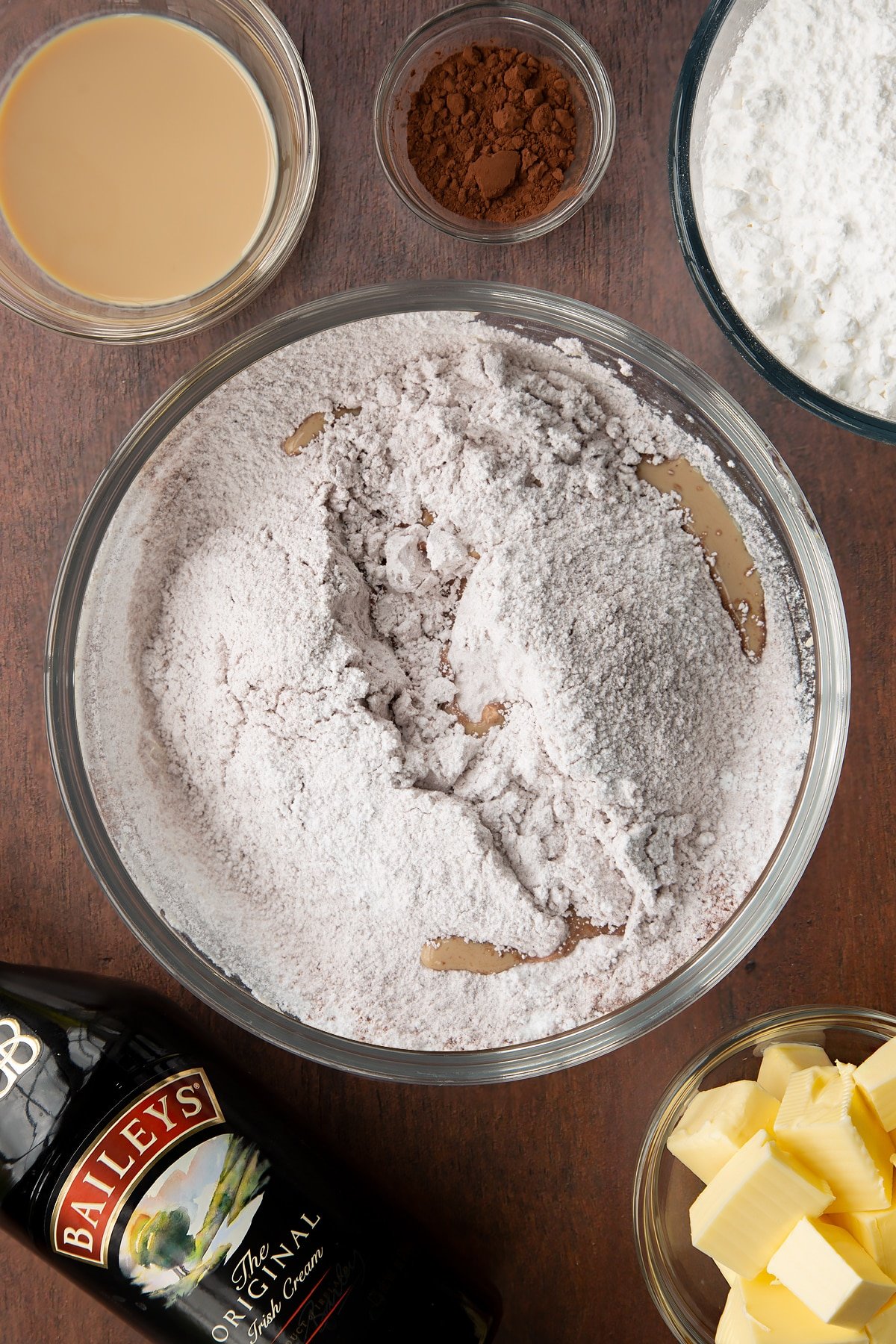 Butter, icing sugar, cocoa and Baileys in a glass mixing bowl. Ingredients to make Baileys chocolate cupcakes surround the bowl.