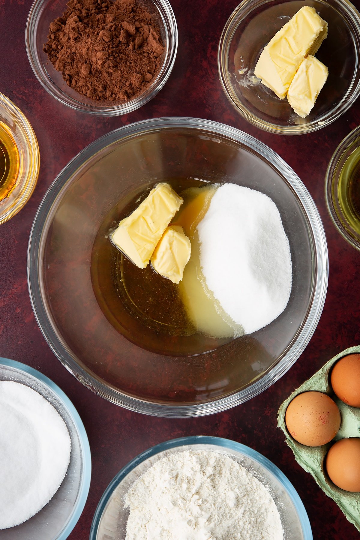 Caster sugar, golden syrup, oil and softened butter in a glass mixing bowl. Ingredients to make chocolate drizzle cake surround the bowl.