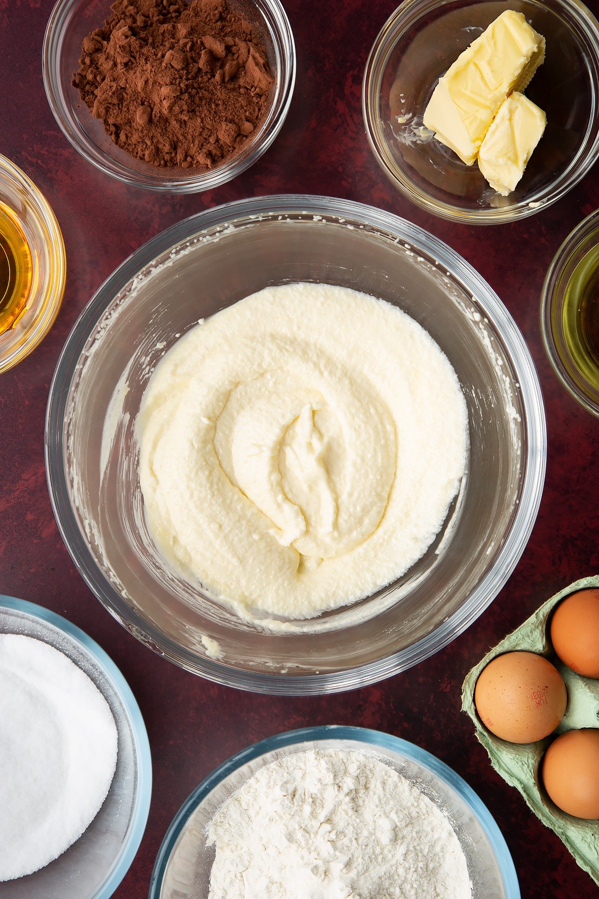 Sugar, golden syrup, oil and butter, whisked together in a glass mixing bowl. Ingredients to make chocolate drizzle cake surround the bowl.