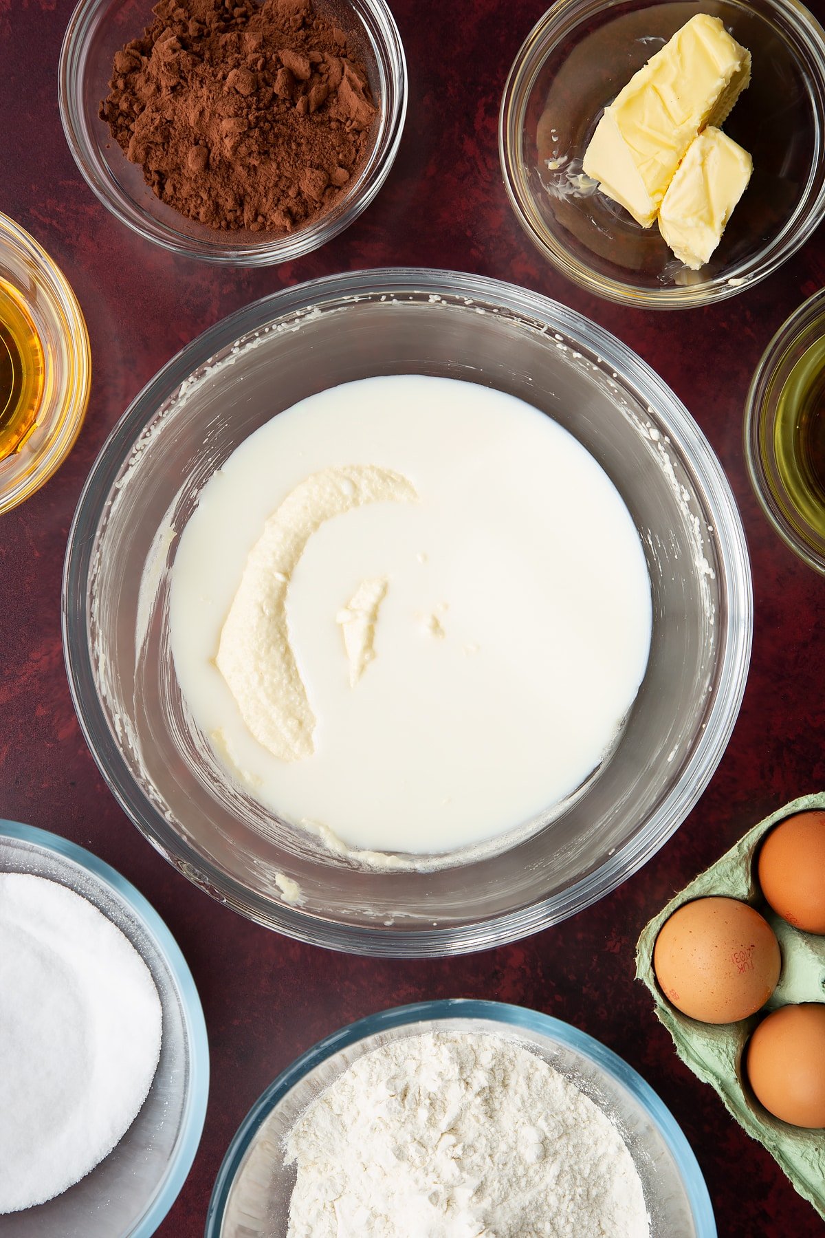 Sugar, golden syrup, oil and butter, whisked together in a glass mixing bowl with milk on top. Ingredients to make chocolate drizzle cake surround the bowl.