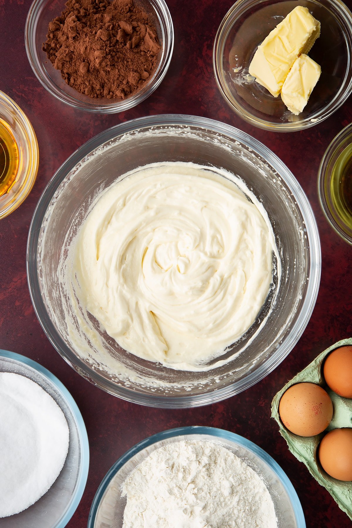 Sugar, golden syrup, oil, milk and butter, whisked together in a glass mixing bowl. Ingredients to make chocolate drizzle cake surround the bowl.