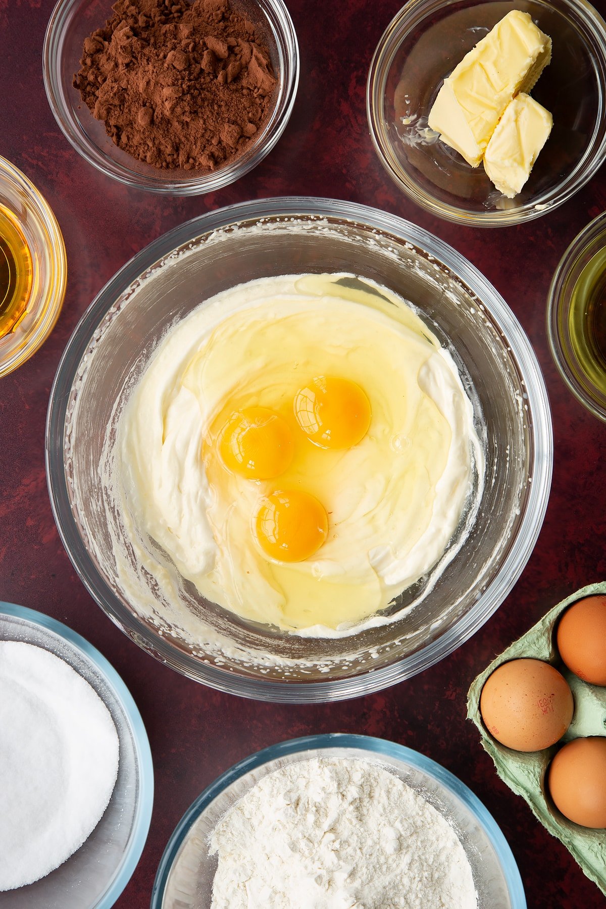 Sugar, golden syrup, oil, milk and butter, whisked together in a glass mixing bowl with eggs on top. Ingredients to make chocolate drizzle cake surround the bowl.