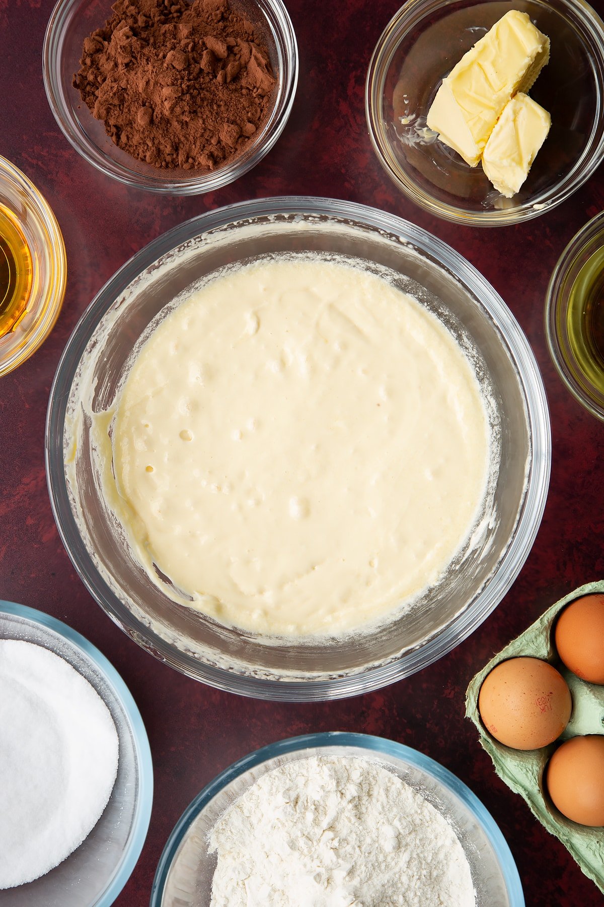 Sugar, golden syrup, oil, milk, eggs and butter, whisked together in a glass mixing bowl. Ingredients to make chocolate drizzle cake surround the bowl.