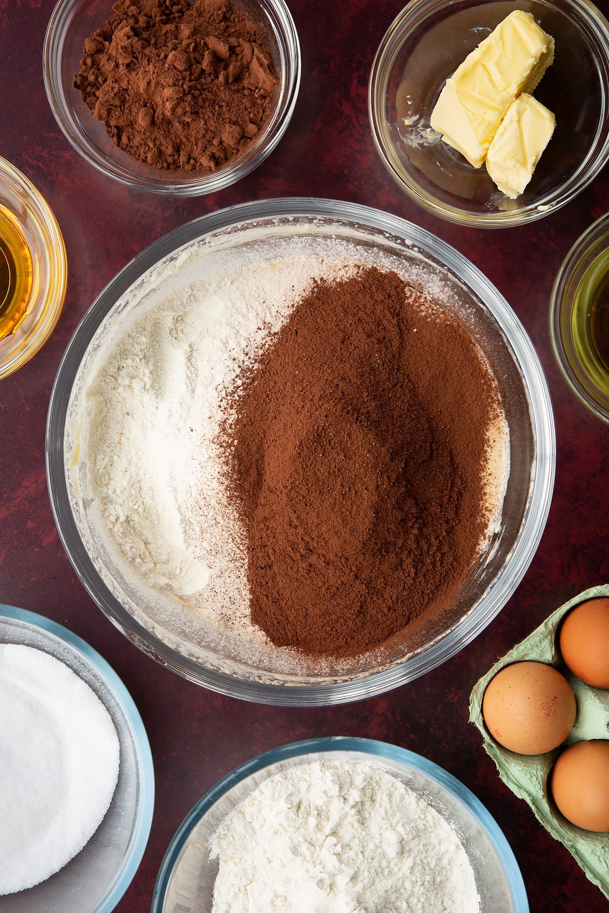 Sugar, golden syrup, oil, milk, eggs and butter, whisked together in a glass mixing bowl with cocoa and flour on top. Ingredients to make chocolate drizzle cake surround the bowl.