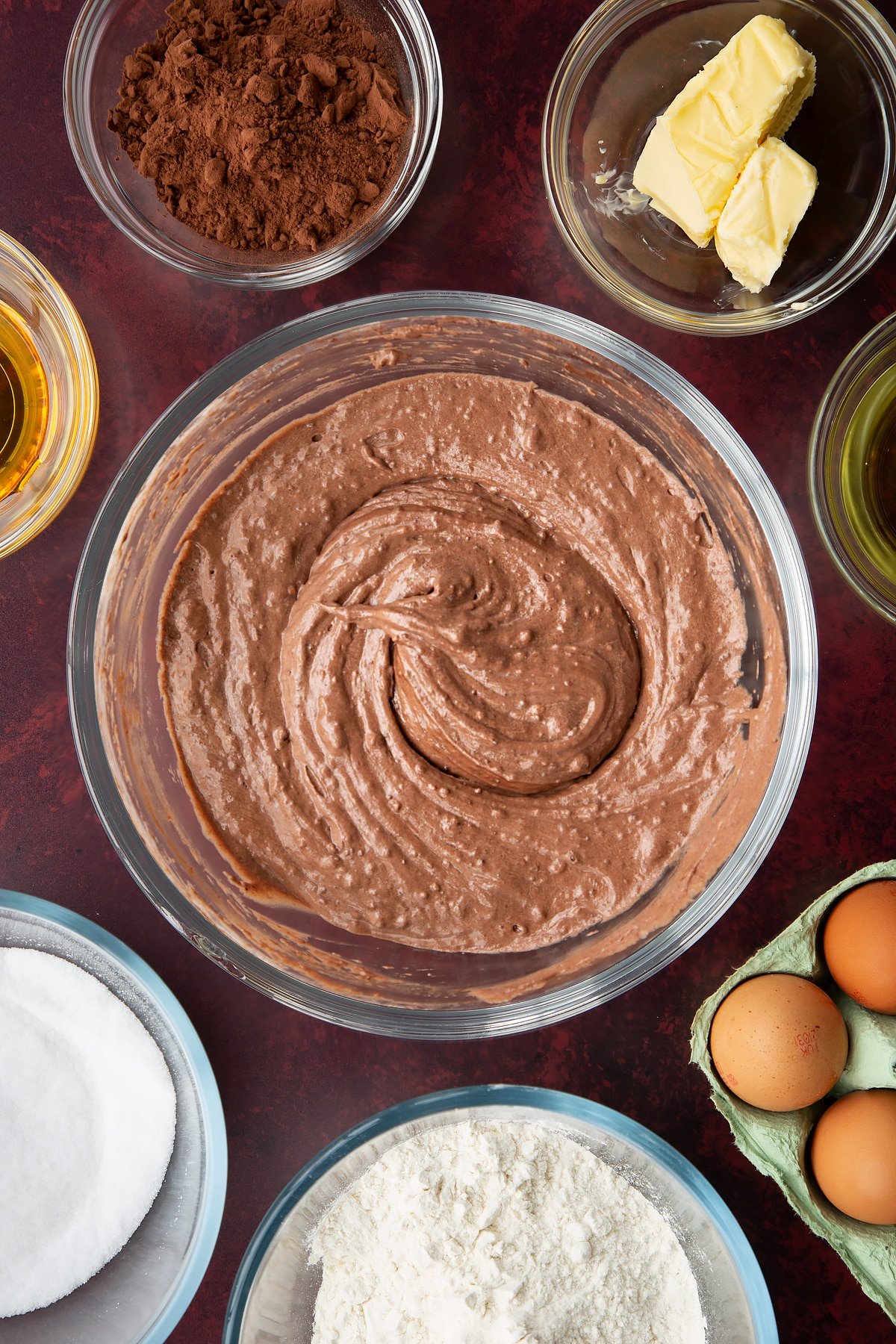 Chocolate drizzle cake batter in a glass mixing bowl. Ingredients to make chocolate drizzle cake surround the bowl.