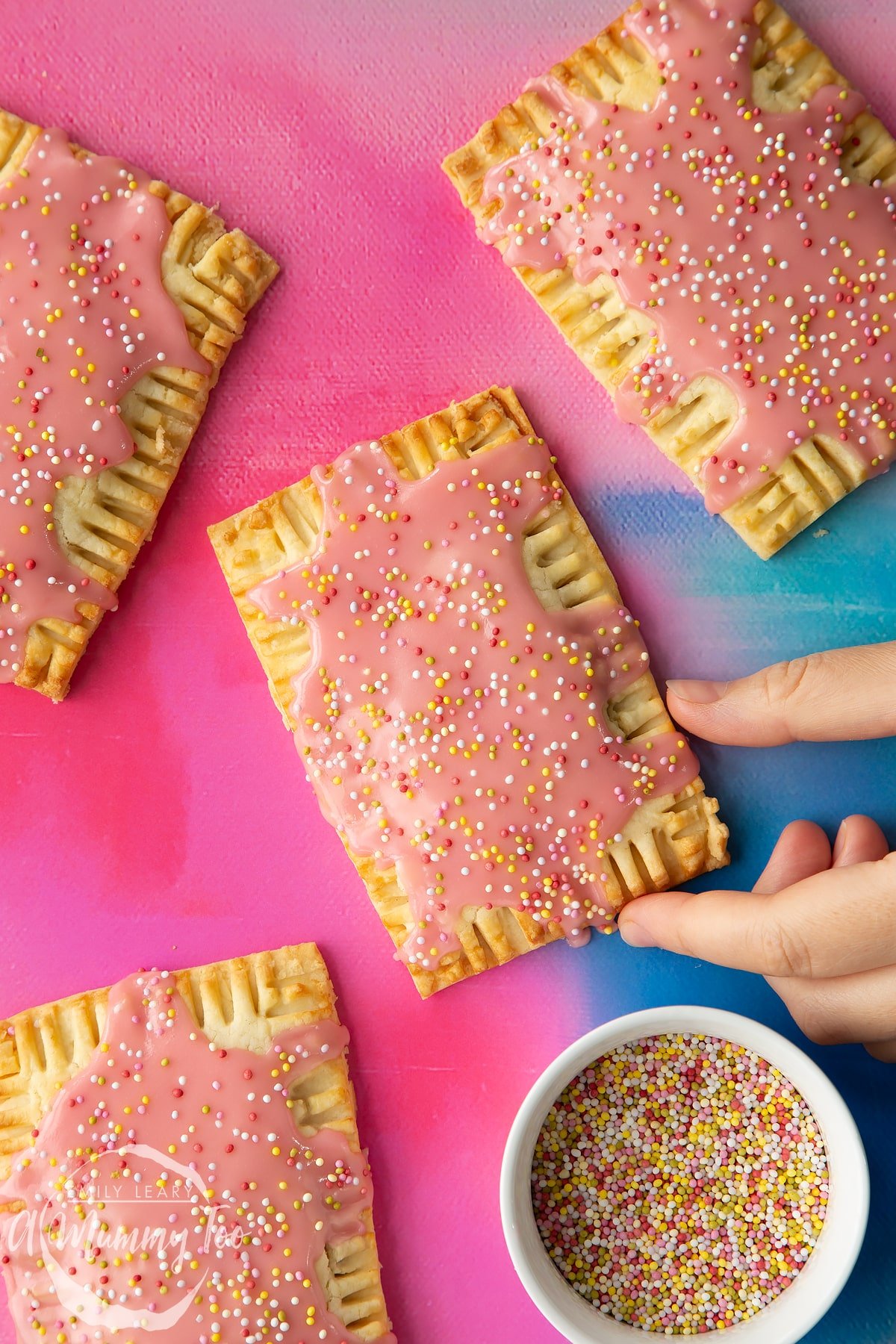 Gluten free pop tarts with pink icing and pastel sprinkles on a bright pink and blue backdrop. A hand reaches for one.