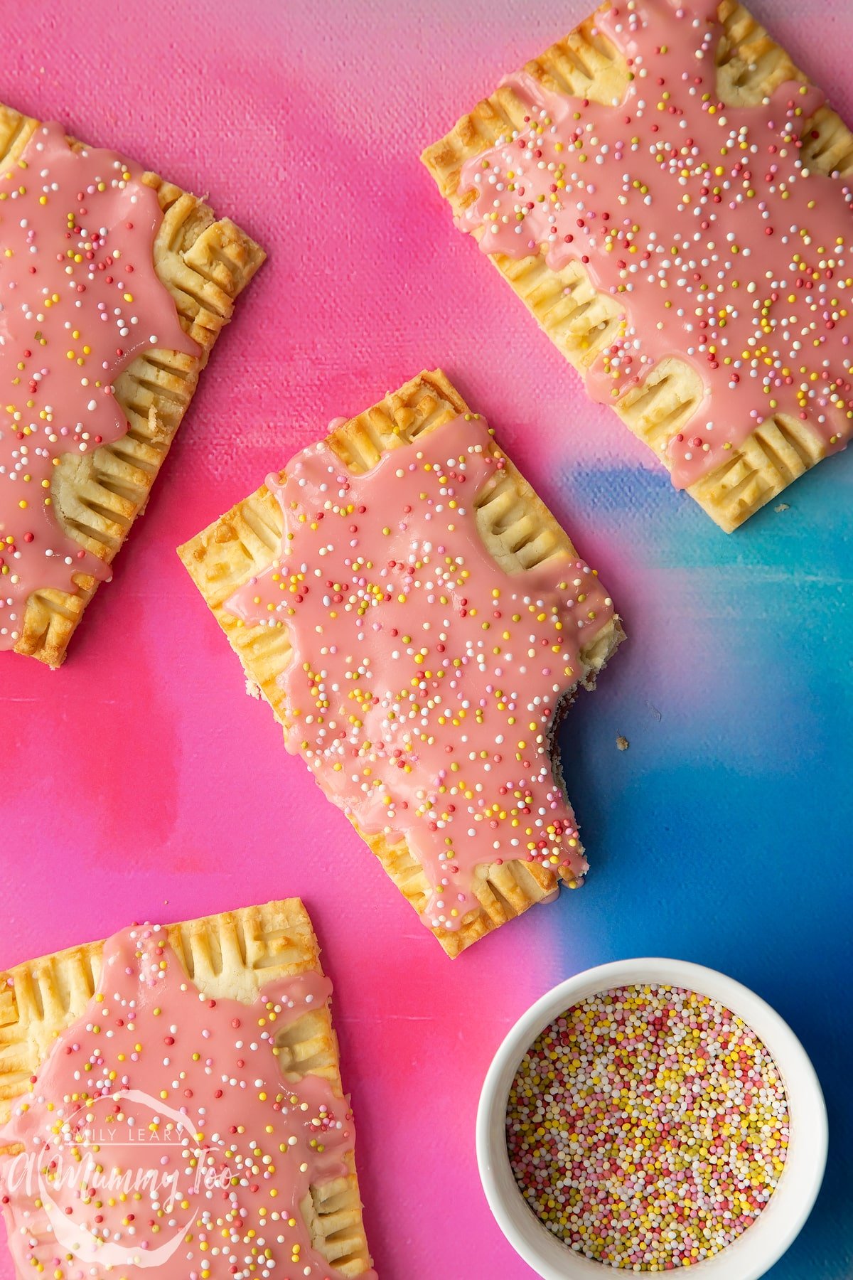 Gluten free pop tarts with pink icing and pastel sprinkles on a bright pink and blue backdrop. One has a bite out of the corner.