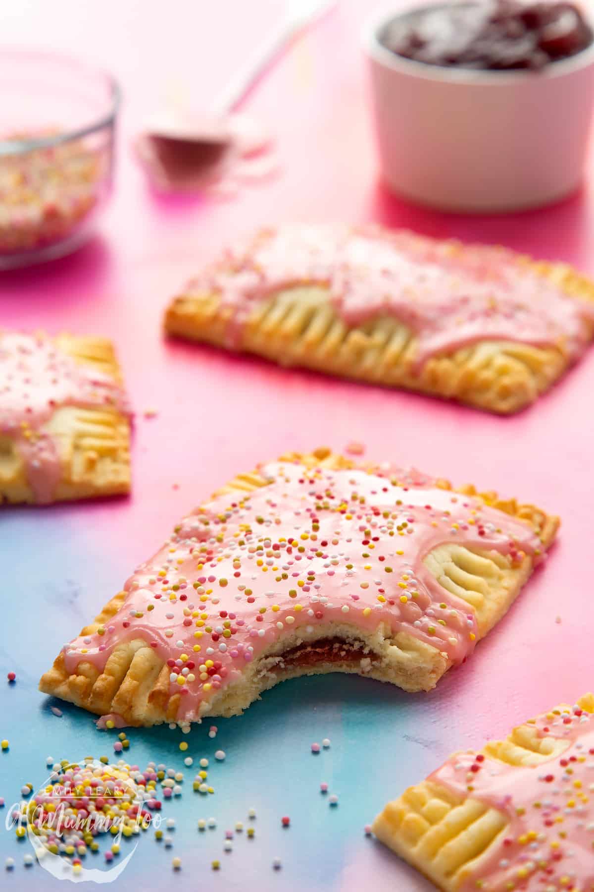 A gluten free pop tart with pink icing and pastel sprinkles. It has a bite out of it. 