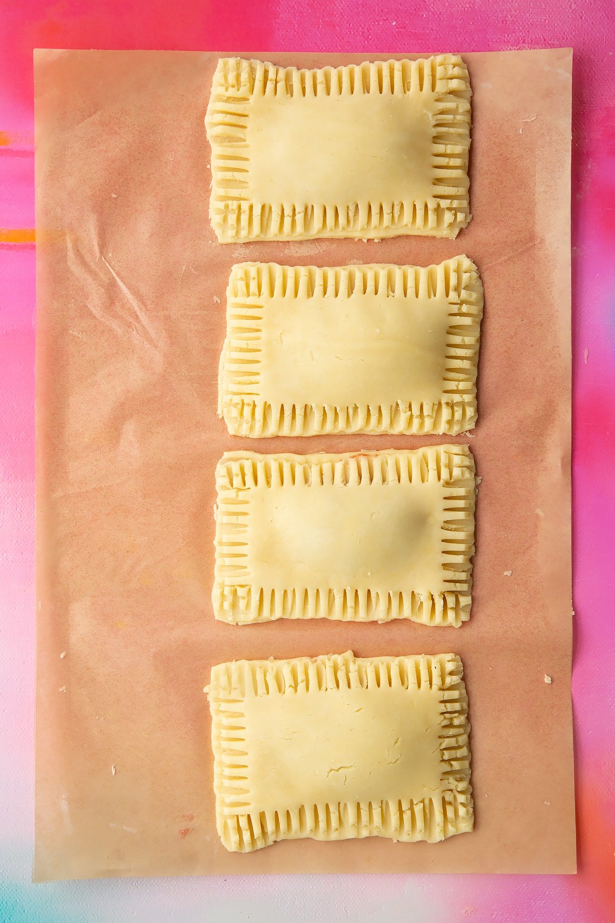 Four raw gluten free pop tarts on baking paper. The edges have been crimped with a fork.