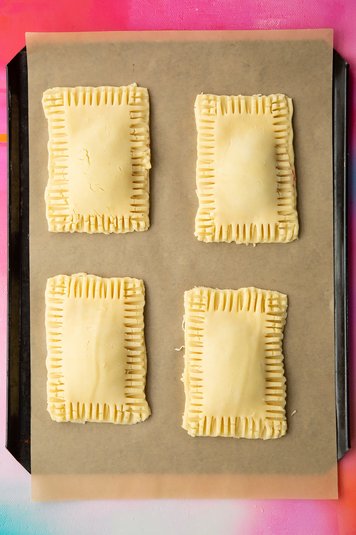 Four raw gluten free pop tarts on baking tray lined with baking paper. The edges have been crimped with a fork.