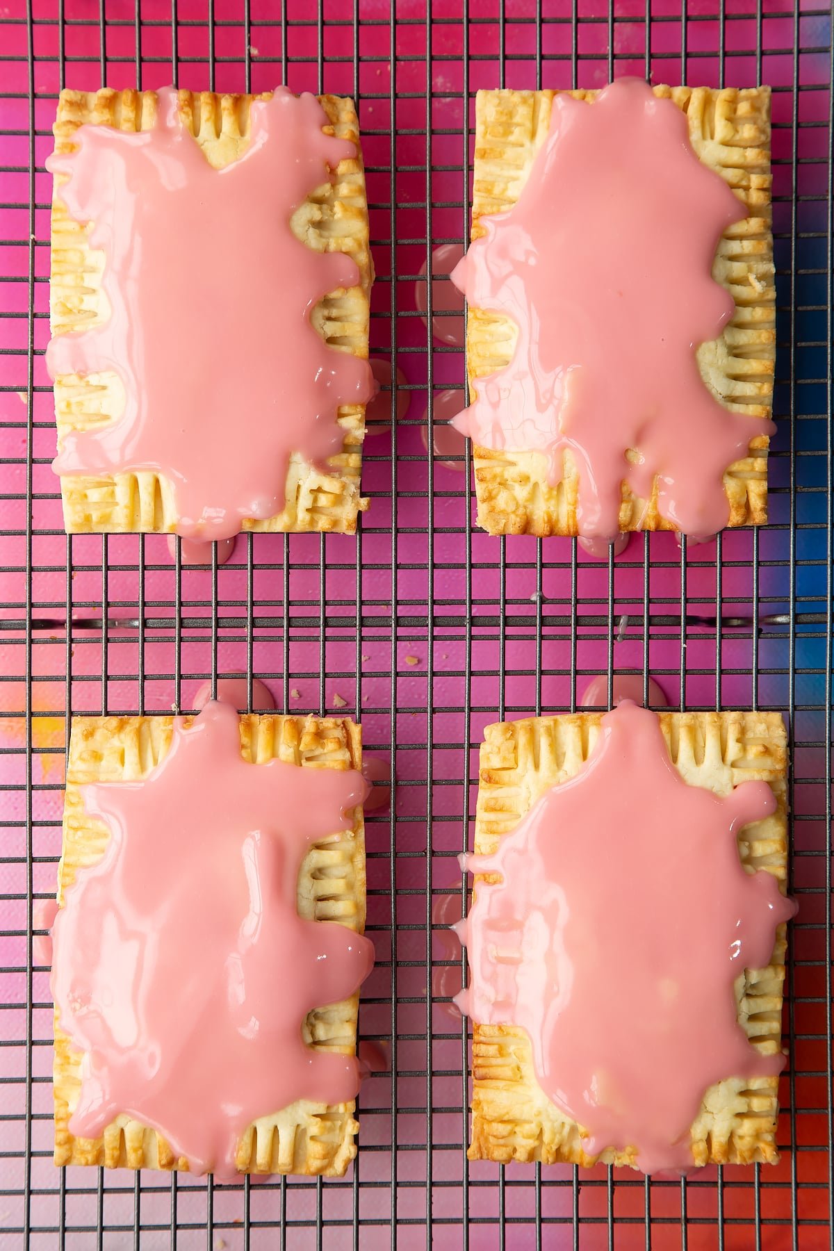 Four freshly baked gluten free pop tarts on a wire cooling rack. They have been drizzled with pink icing.