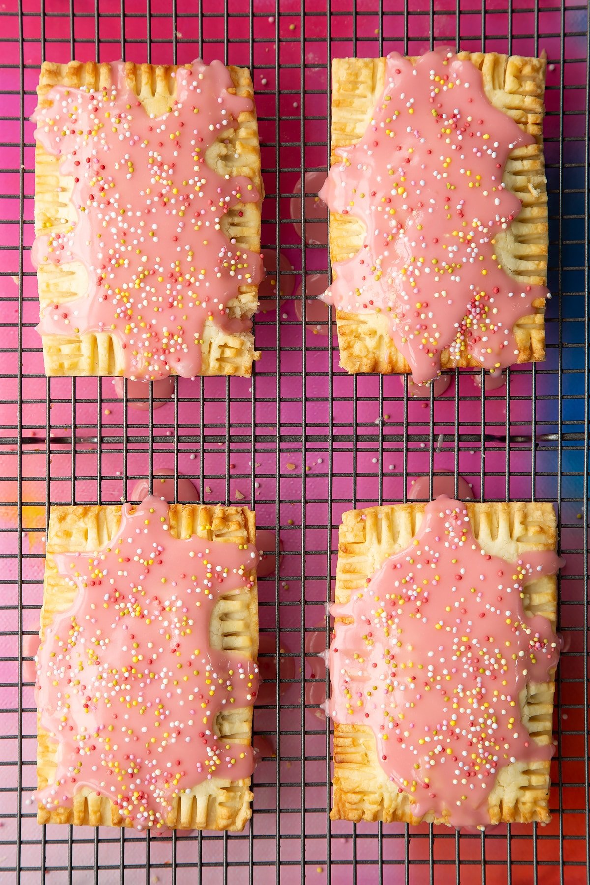Four freshly baked gluten free pop tarts on a wire cooling rack. They have been drizzled with pink icing and covered with sprinkles.