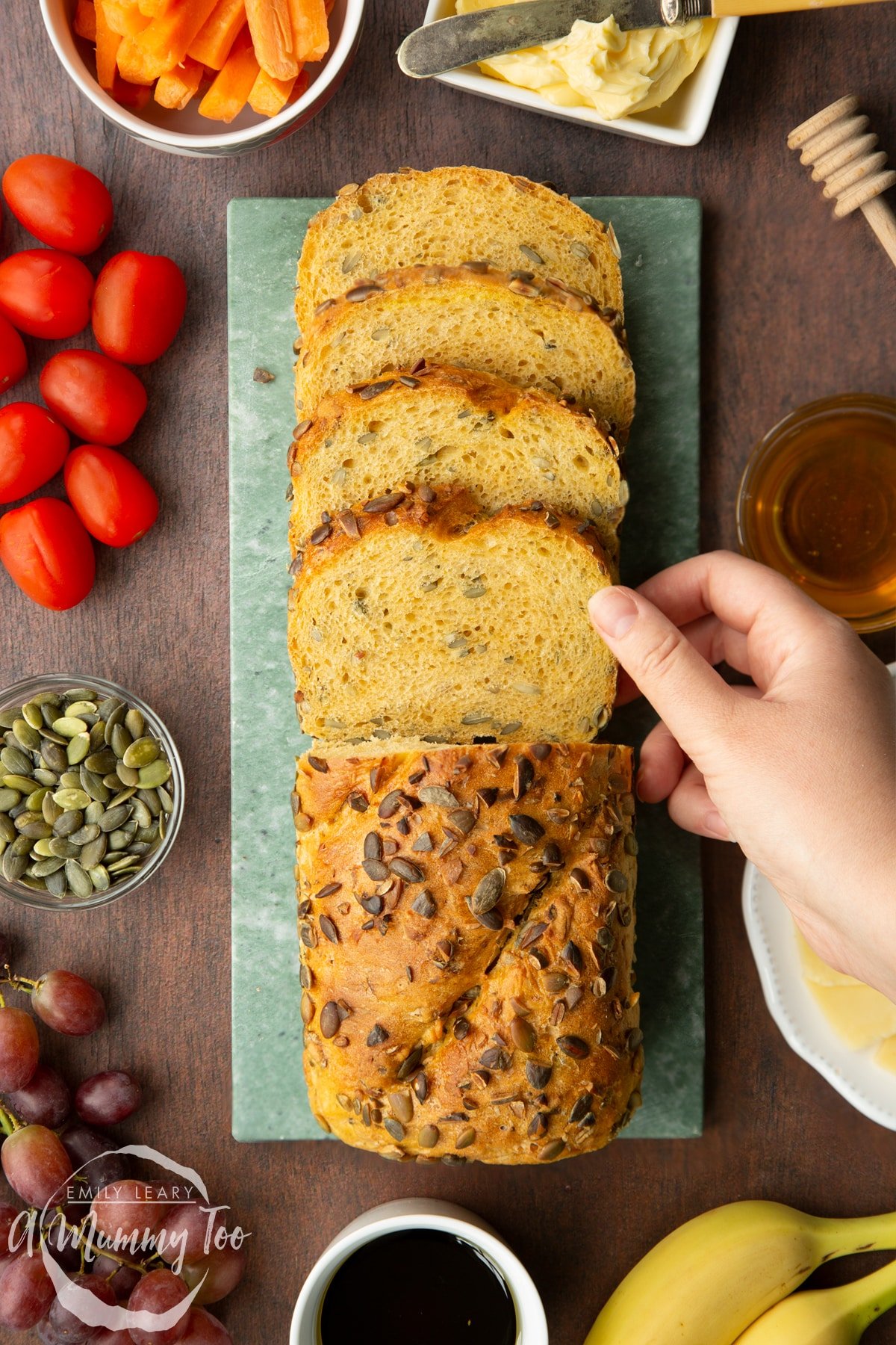 A pumpkin seed bread loaf on a marble board. Half of the loaf has been cut into slices. A hand reaches for a slice.