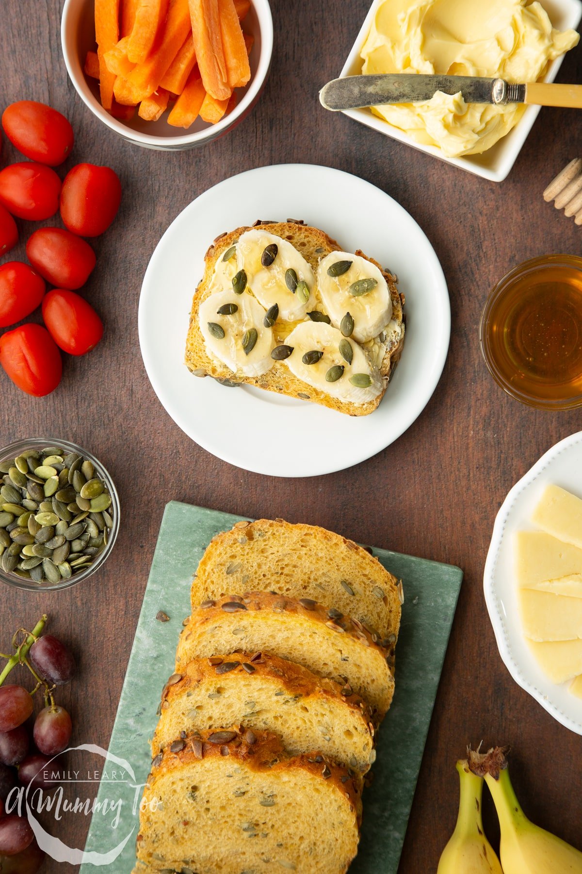 A buttered slice of pumpkin seed bread topped with sliced banana, pumpkin seeds and honey on a white plate. More slices are on a marble board.