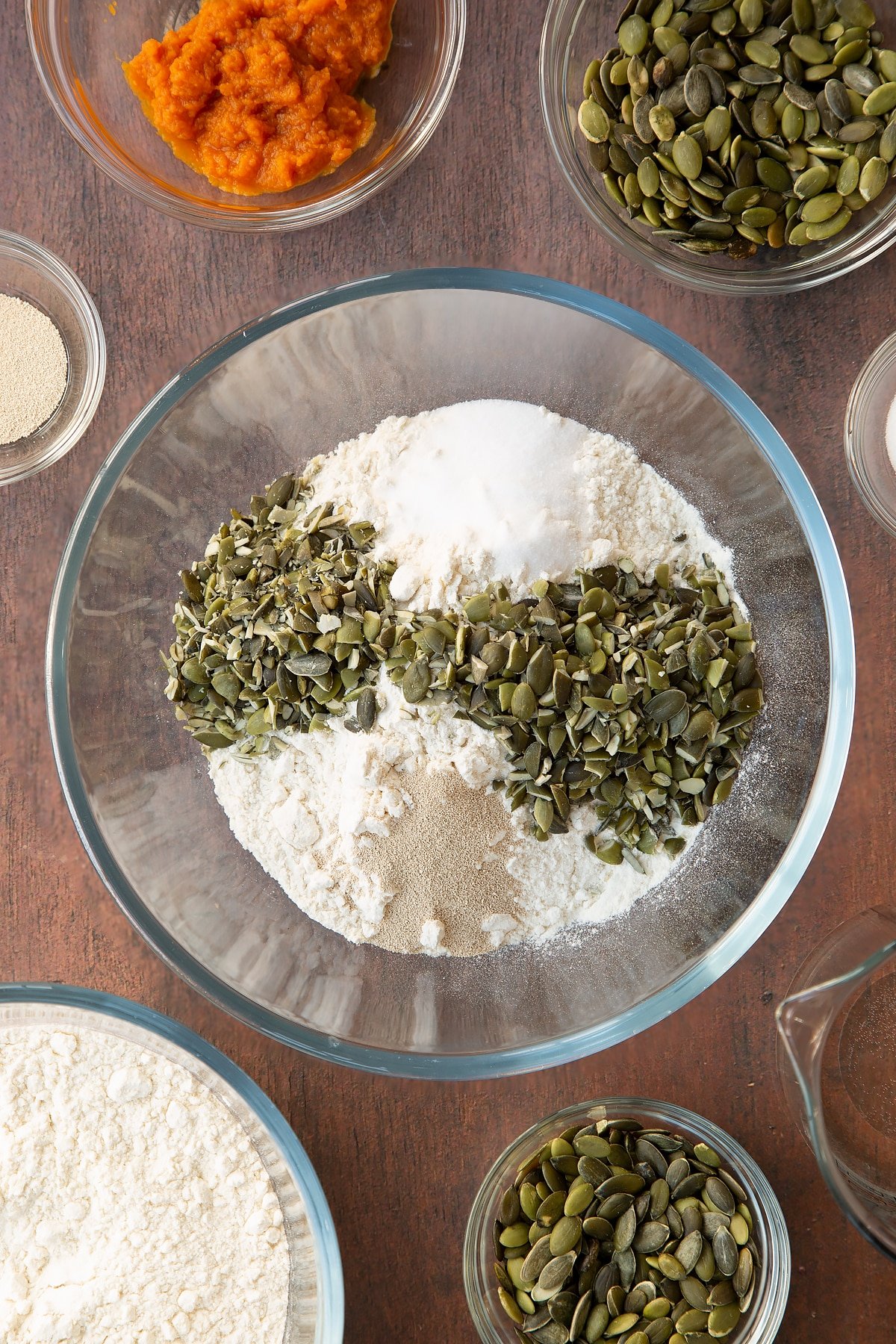 Flour, salt, yeast and pumpkin seeds in a glass mixing bowl. Ingredients to make a pumpkin seed bread recipe surround the bowl. 