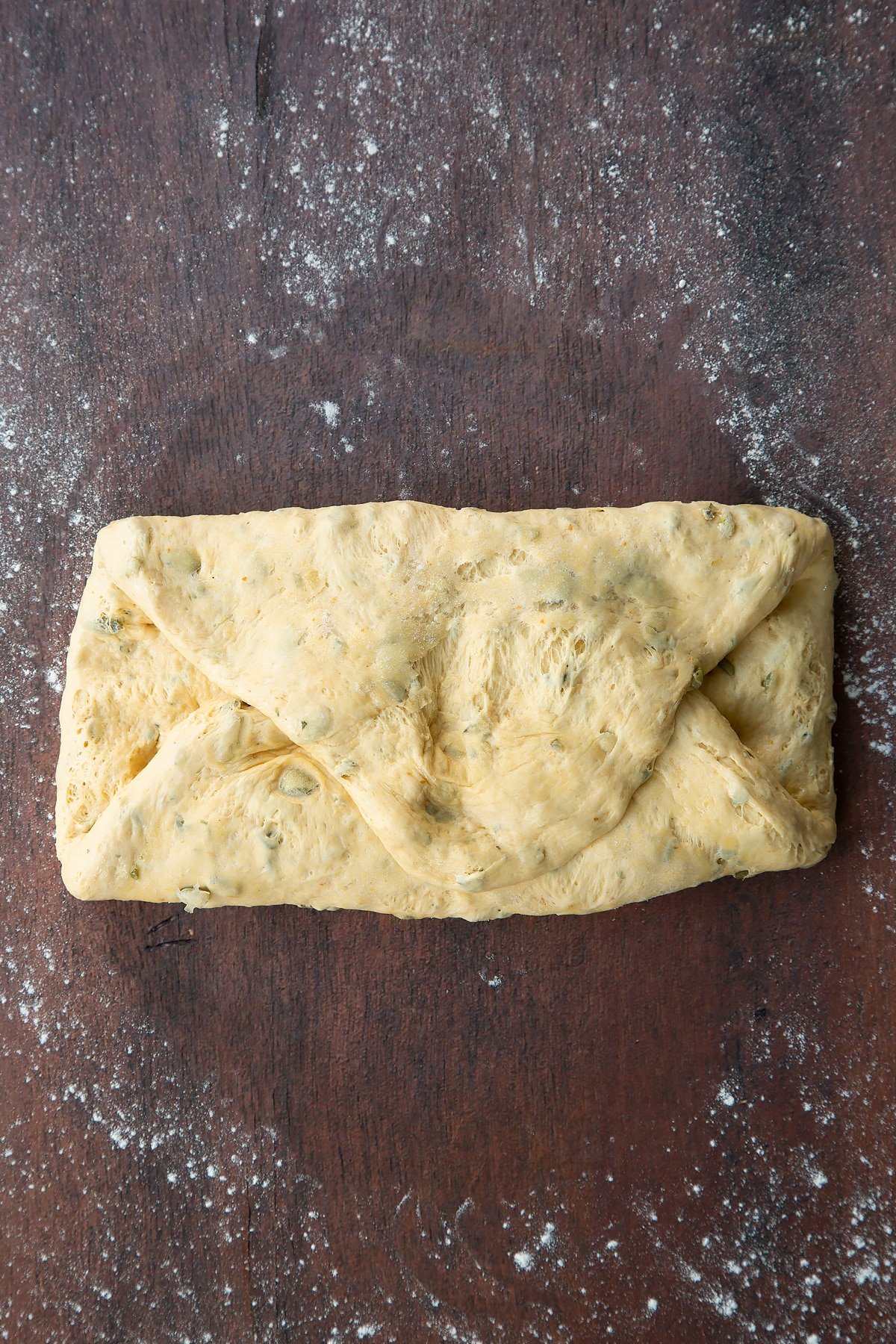 Pumpkin seed bread dough pressed into a flat oval then folded into three on a floured surface.