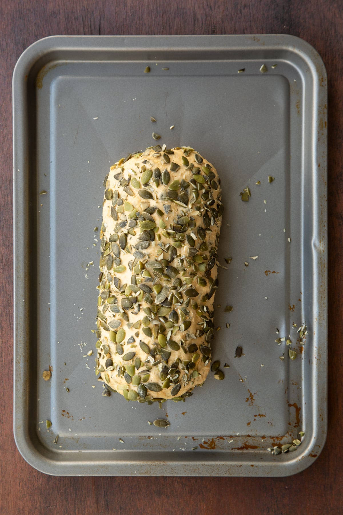 A roll of pumpkin seed bread dough, rolled in pumpkin seeds on a metal tray.