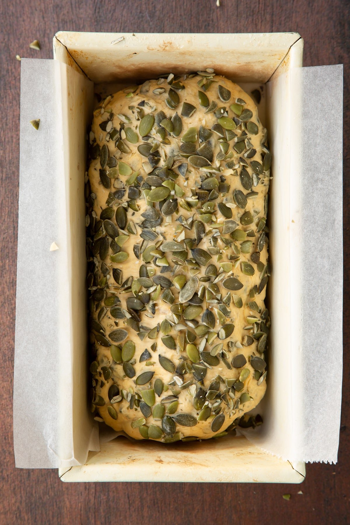 Pumpkin seed specie dough covered with pumpkin seeds in a loaf tin lined with sultry paper.