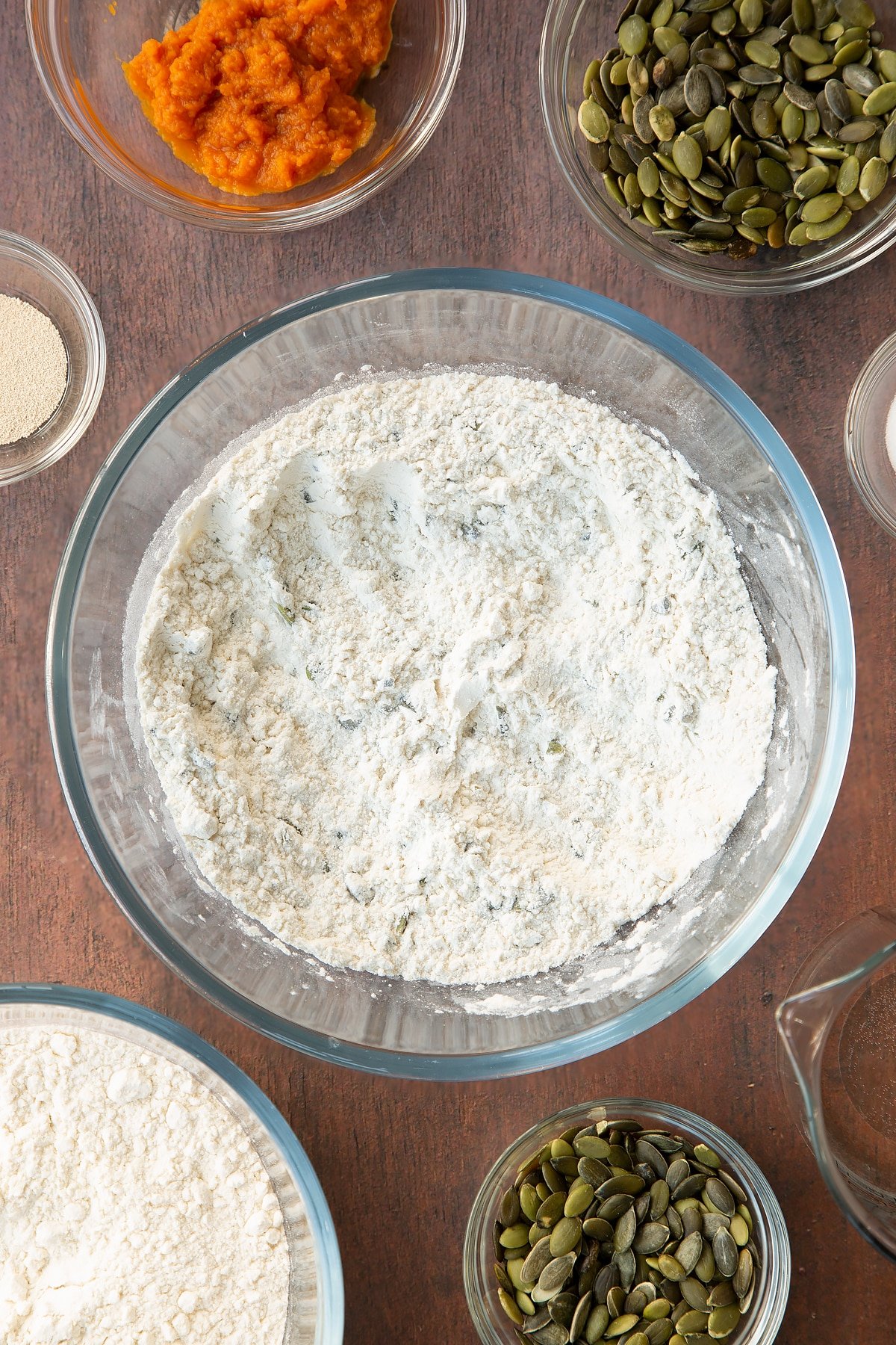 Flour, salt, yeast and pumpkin seeds mixed together in a glass mixing bowl. Ingredients to make a pumpkin seed bread recipe surround the bowl. 