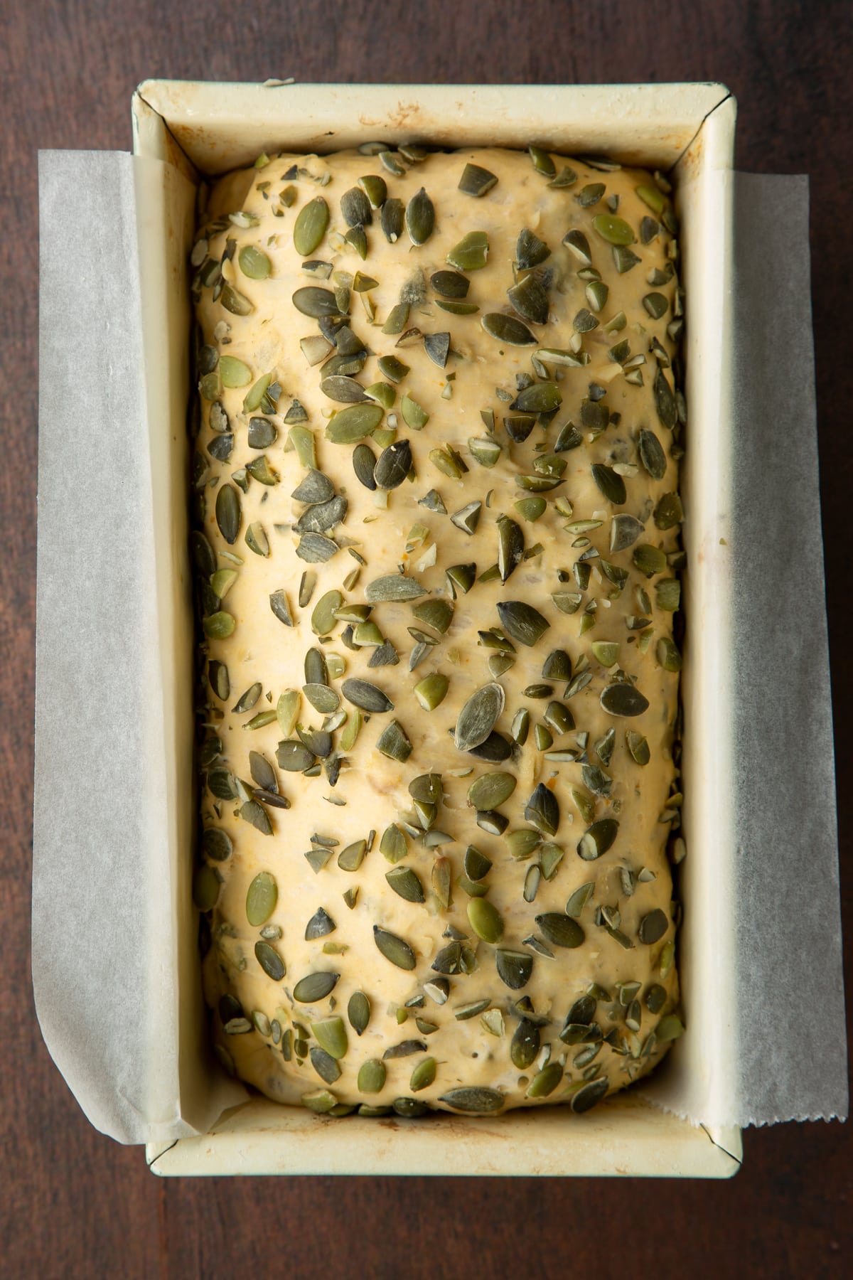 Proved pumpkin seed bread dough covered with pumpkin seeds in a loaf tin lined with baking paper.